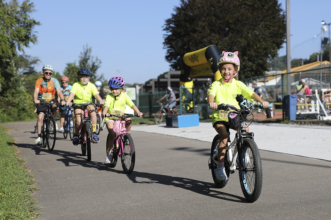 Smiling children and adults in yellow jerseys ride bicycles on a sunny paved trail.