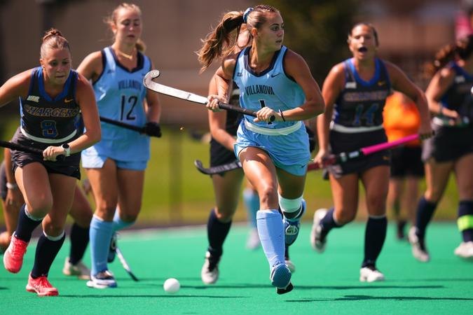 A field hockey player in a light blue uniform runs with a stick on a turf field.