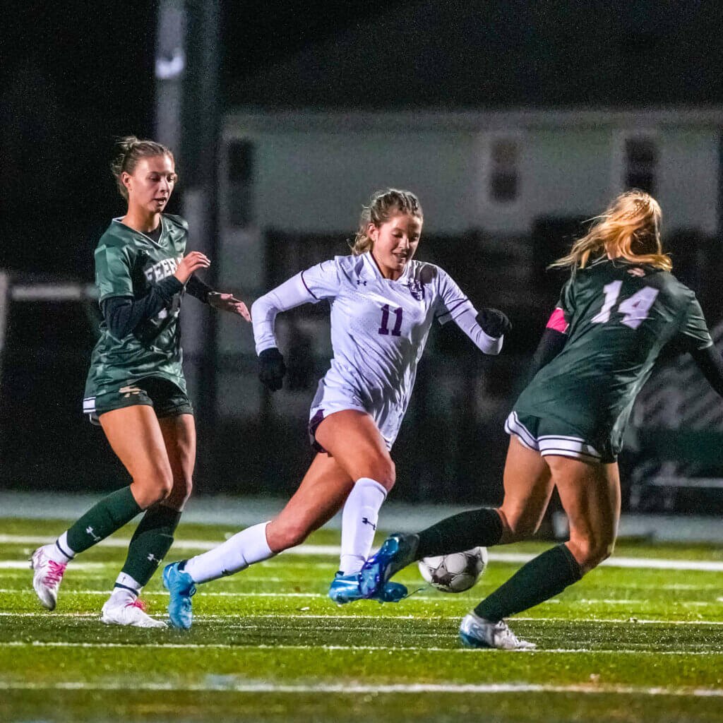 Female soccer player in white jersey number 11 dribbles between two defenders in green jerseys at night.