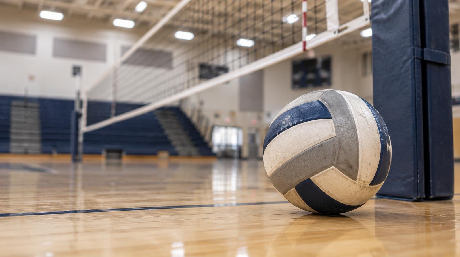 A volleyball resting on a hardwood gymnasium floor near a net post.