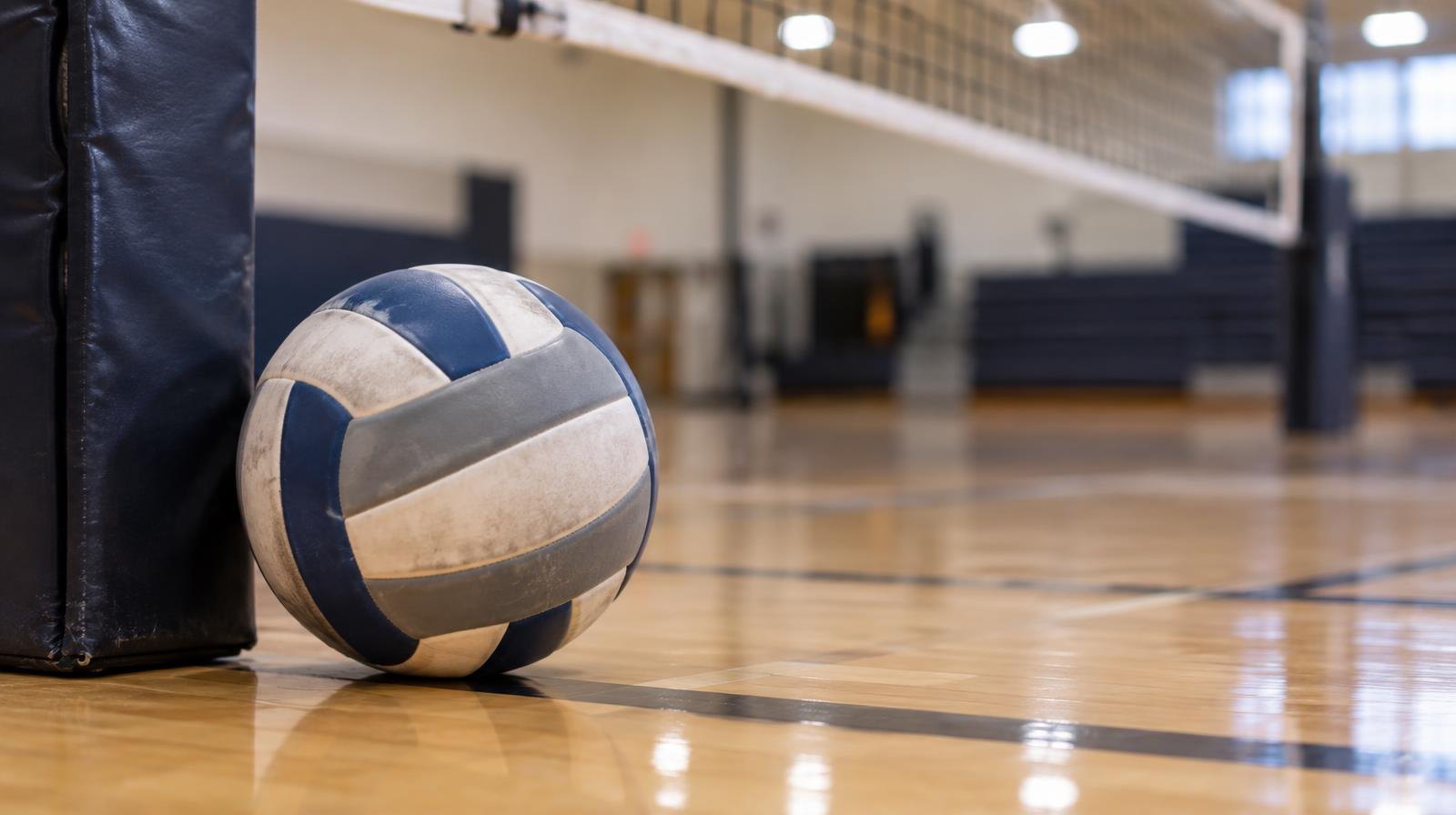 A volleyball rests on a hardwood gymnasium floor beside a net post.