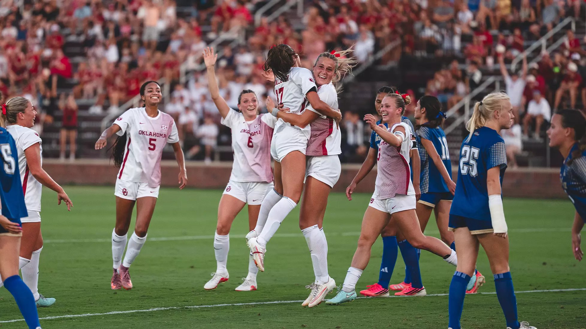 Soccer players in white and crimson uniforms jump and embrace in celebration on the field.