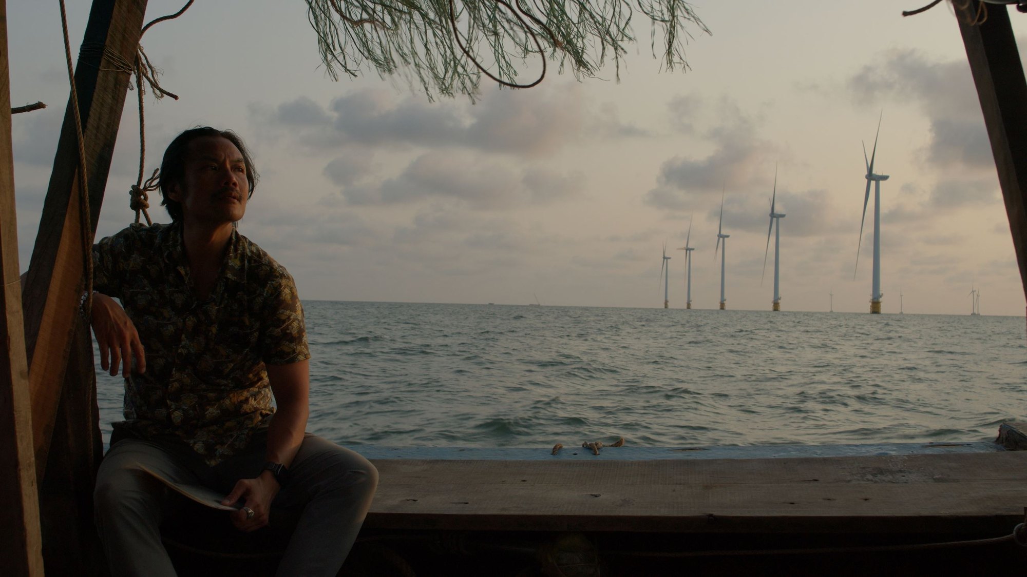 A person in a floral shirt sits on a wooden boat structure gazing toward a calm sea with wind turbines on the horizon.