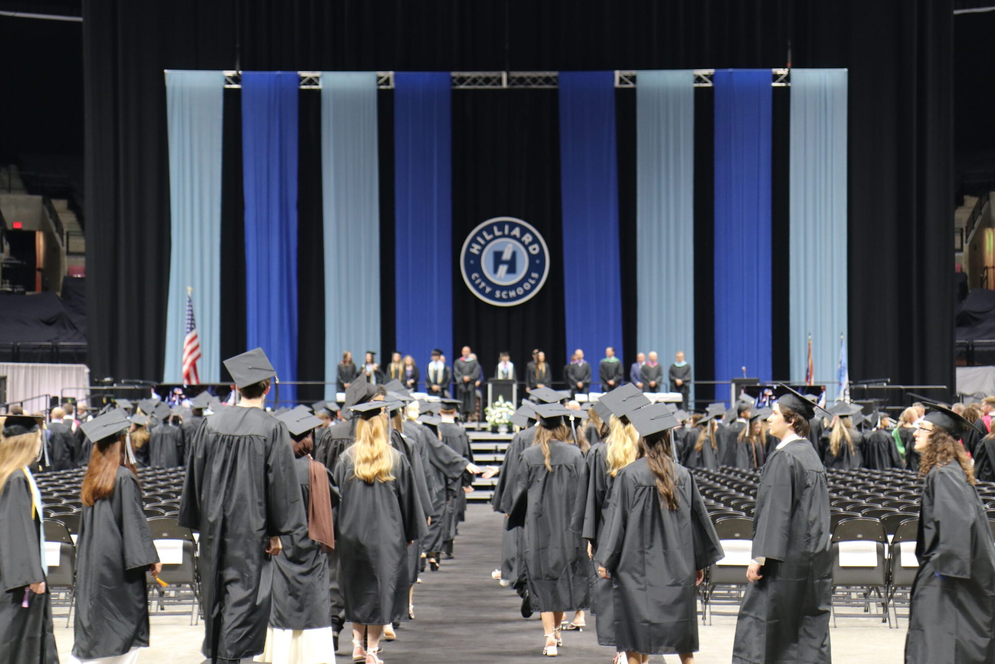 Graduates in black caps and gowns walk toward a decorated stage with a school district seal.