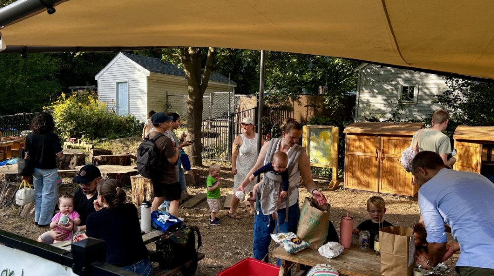 Adults and toddlers gathered under a canopy at an outdoor early childhood play area.