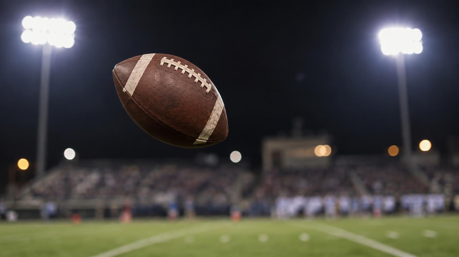An American football spinning through the air above a green grass field under bright stadium lights.