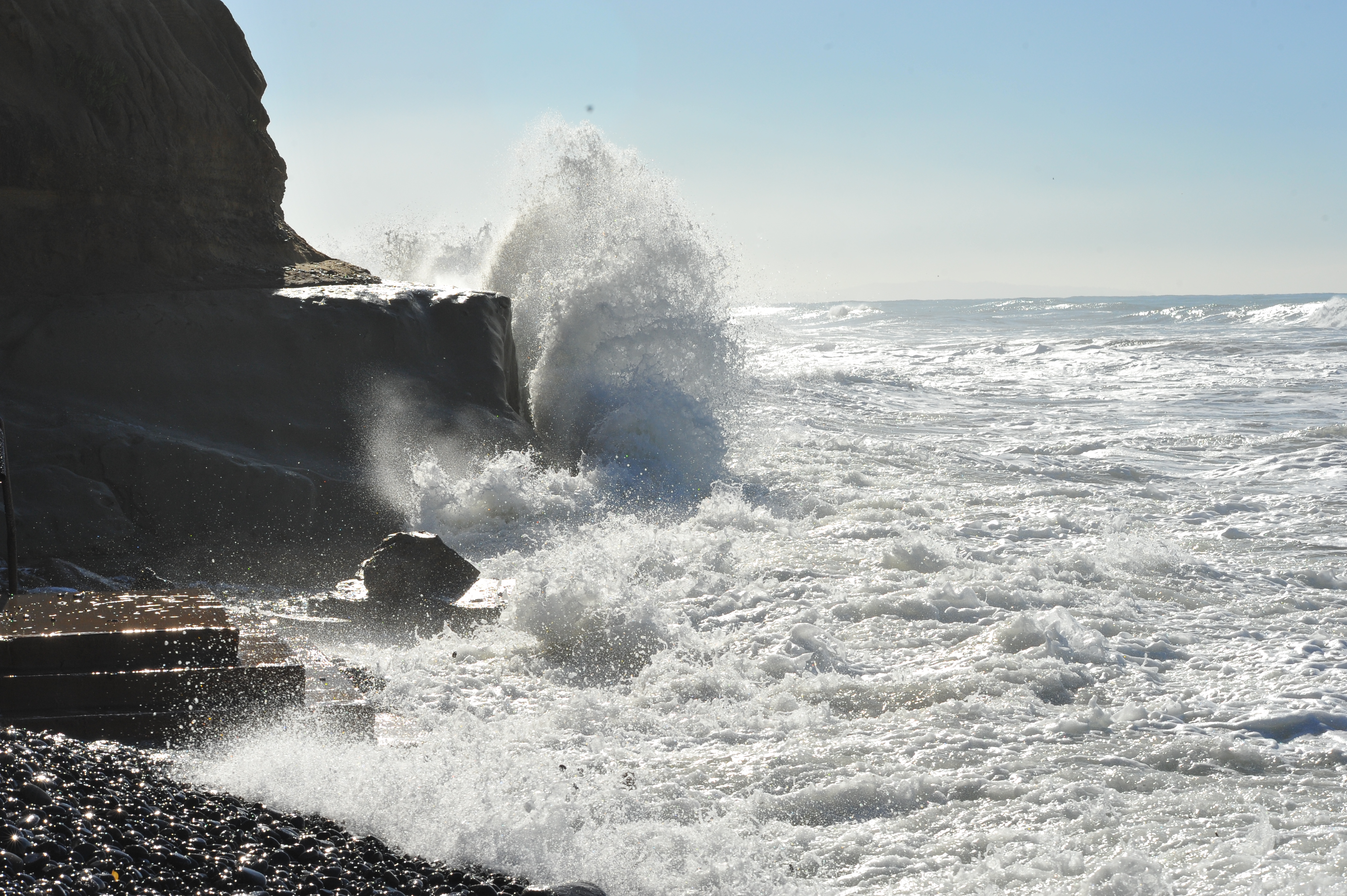 Large waves crashing against a rocky cliff base on a cobblestone beach.