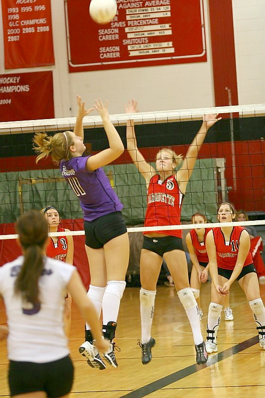 A player in purple spikes the ball over the net as two players in red attempt to block.