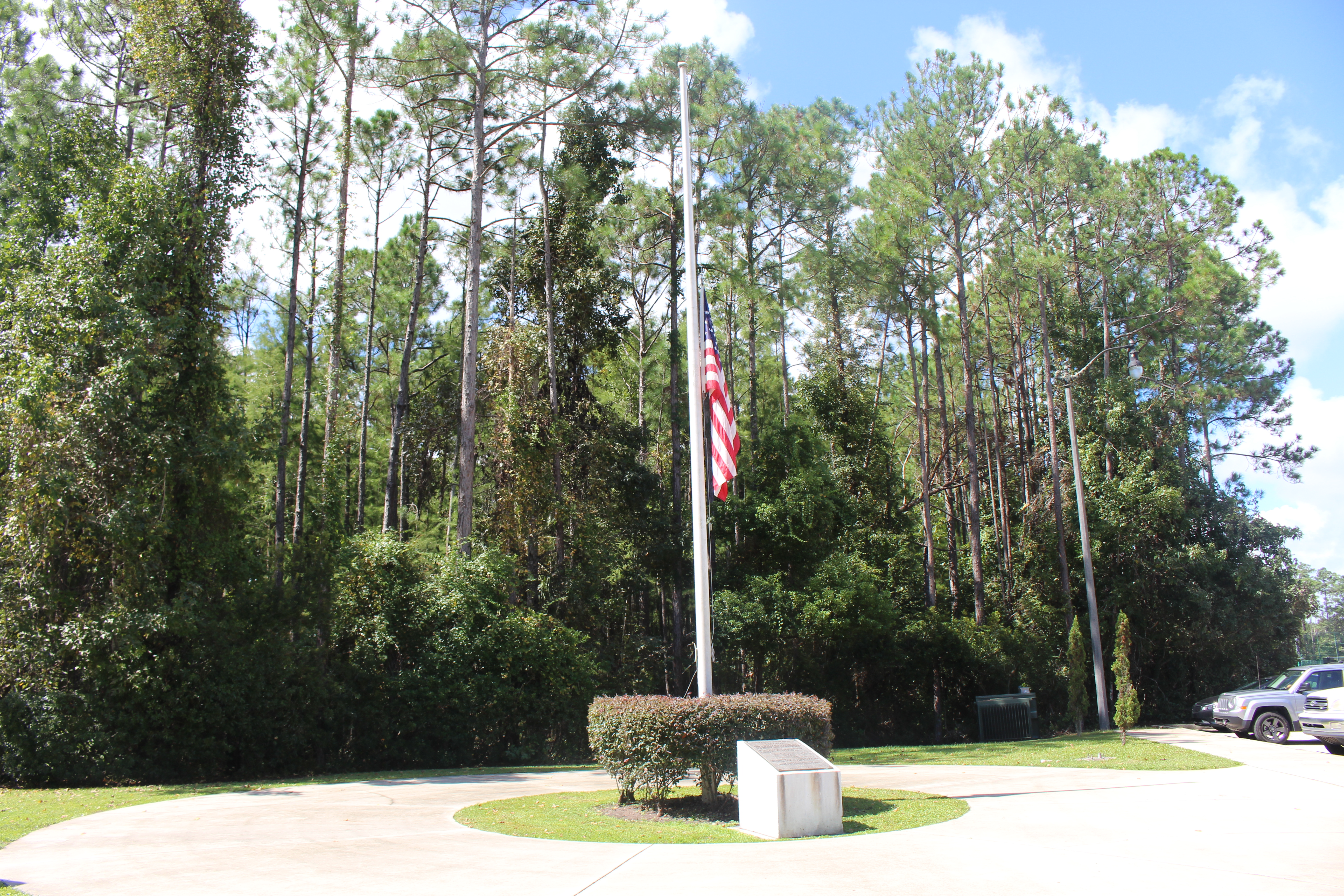 Flagpole with American flag at half staff in a landscaped park surrounded by tall pine trees.
