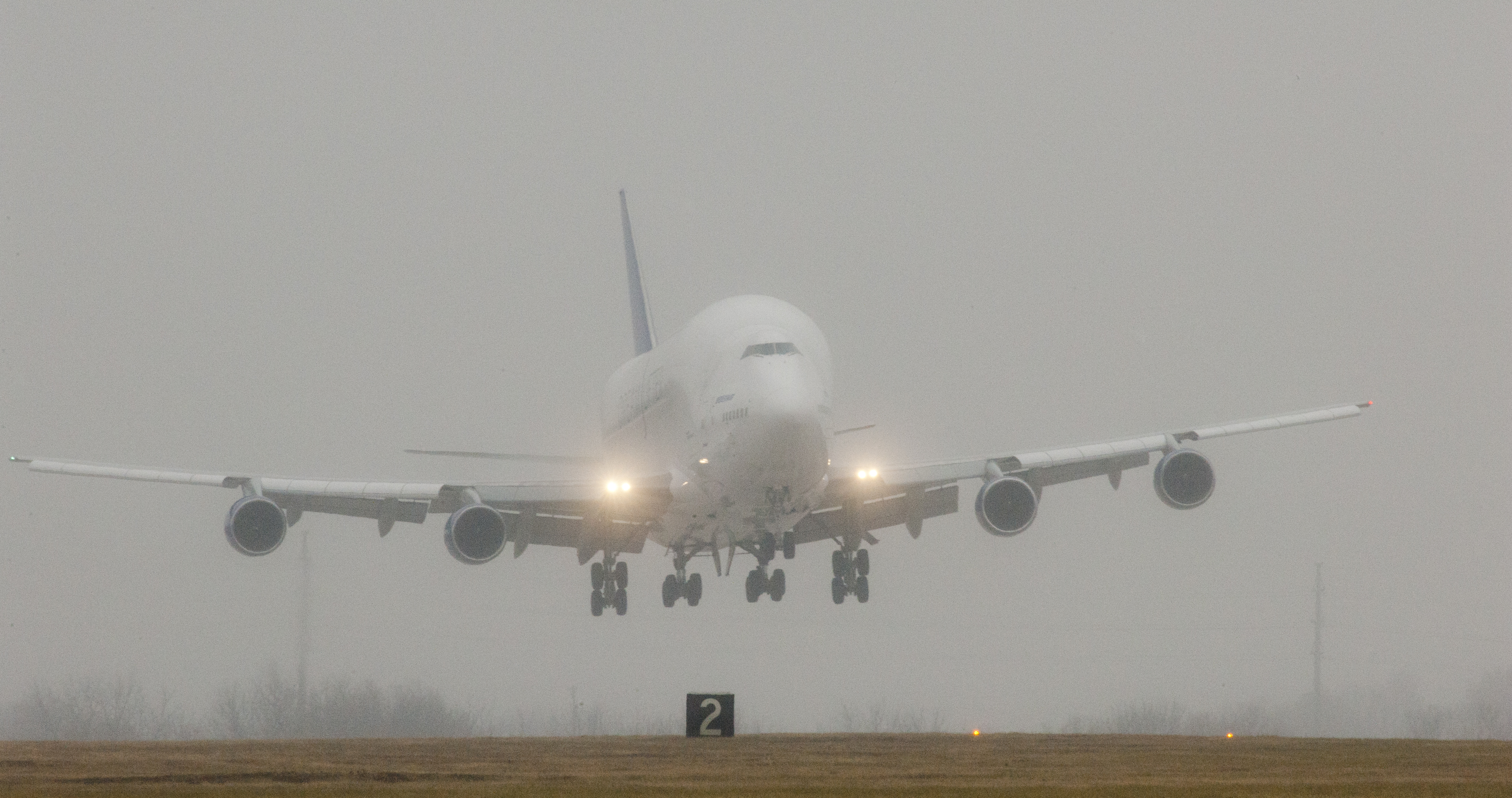 Large white Boeing aircraft with landing gear down touches down on a runway in overcast conditions.