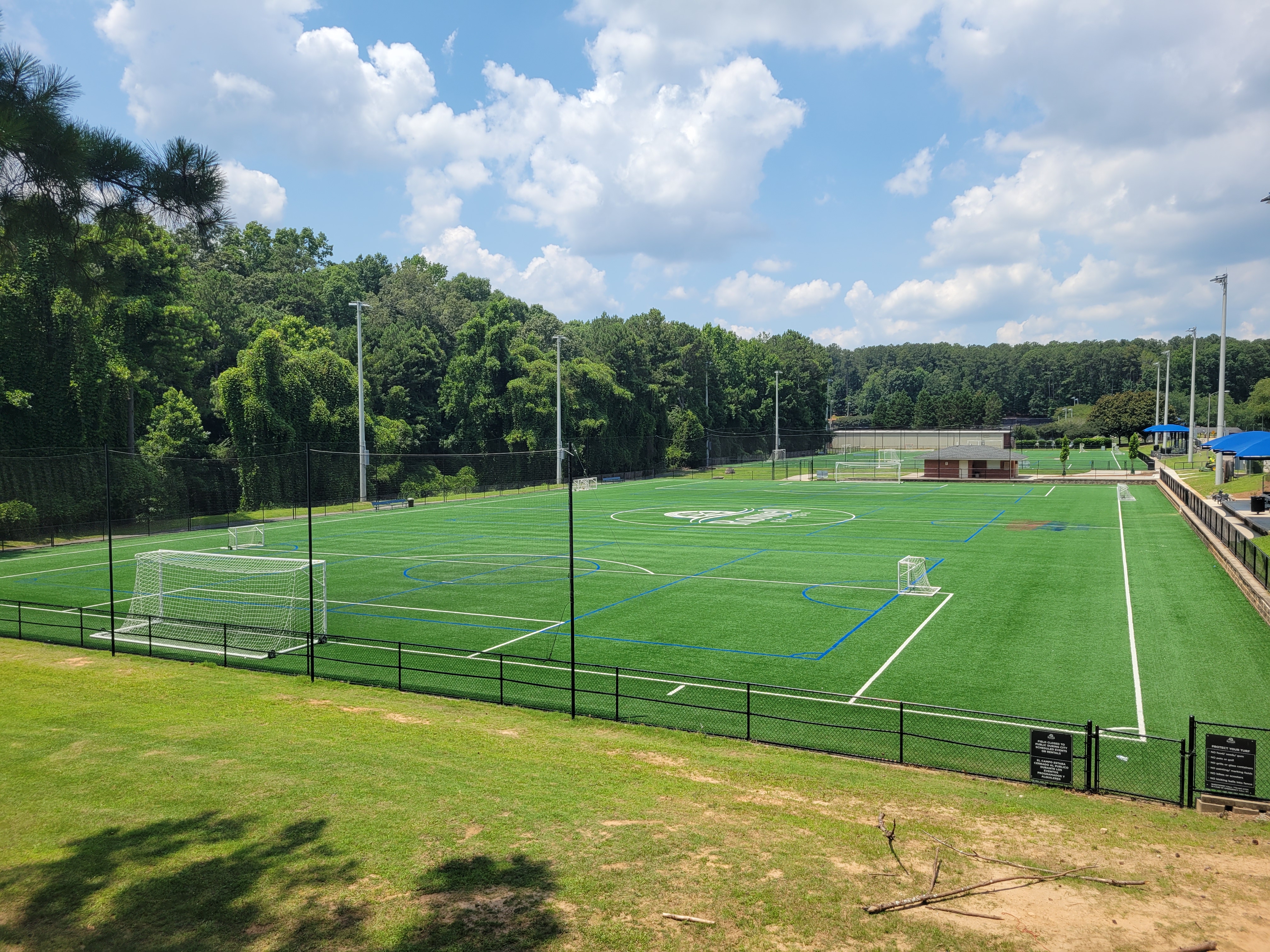 Outdoor artificial turf athletic fields with tall light poles and chain-link fencing at a public park.