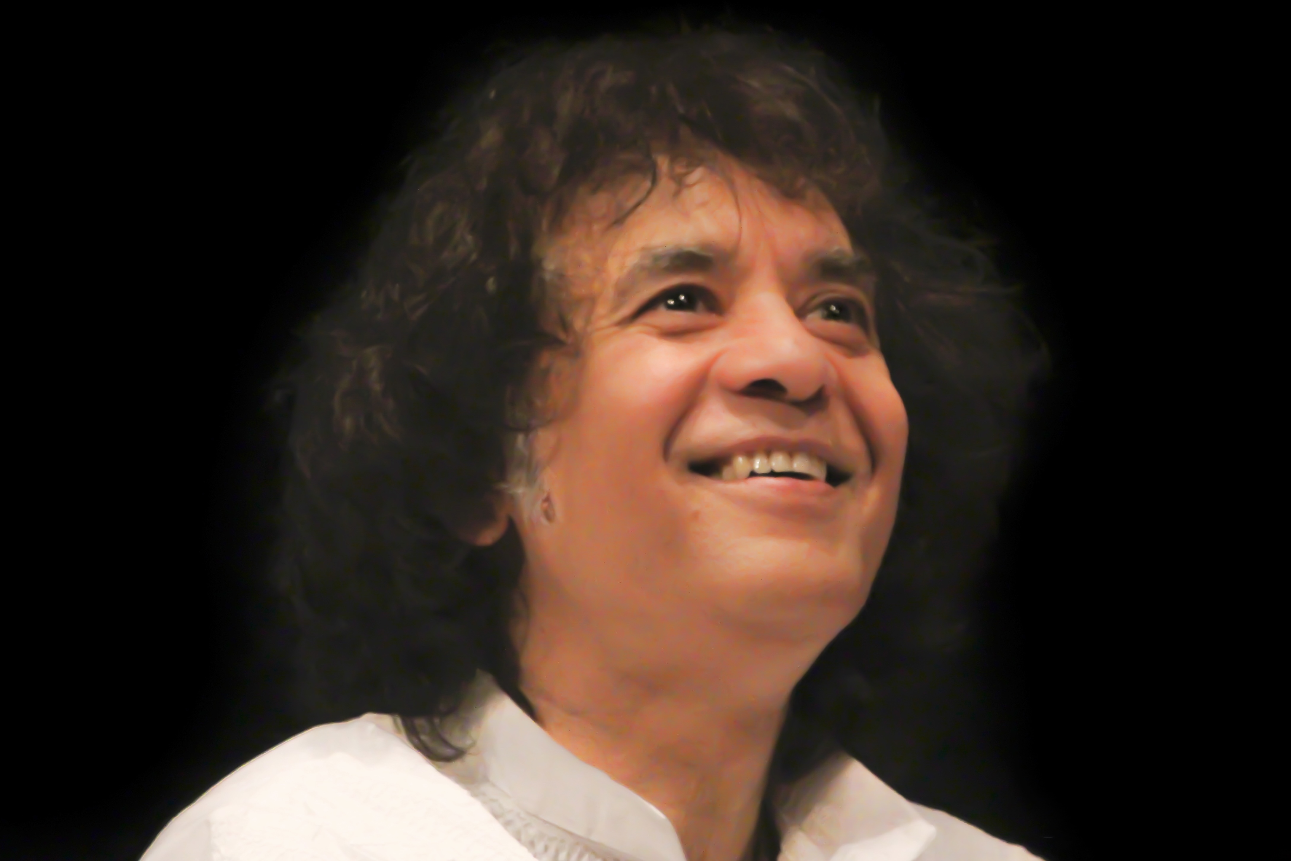 A man with curly dark hair wearing a white shirt smiles against a dark background.