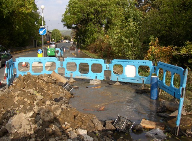 Excavated road trench with blue safety barriers and water pooling around a cut pipe.