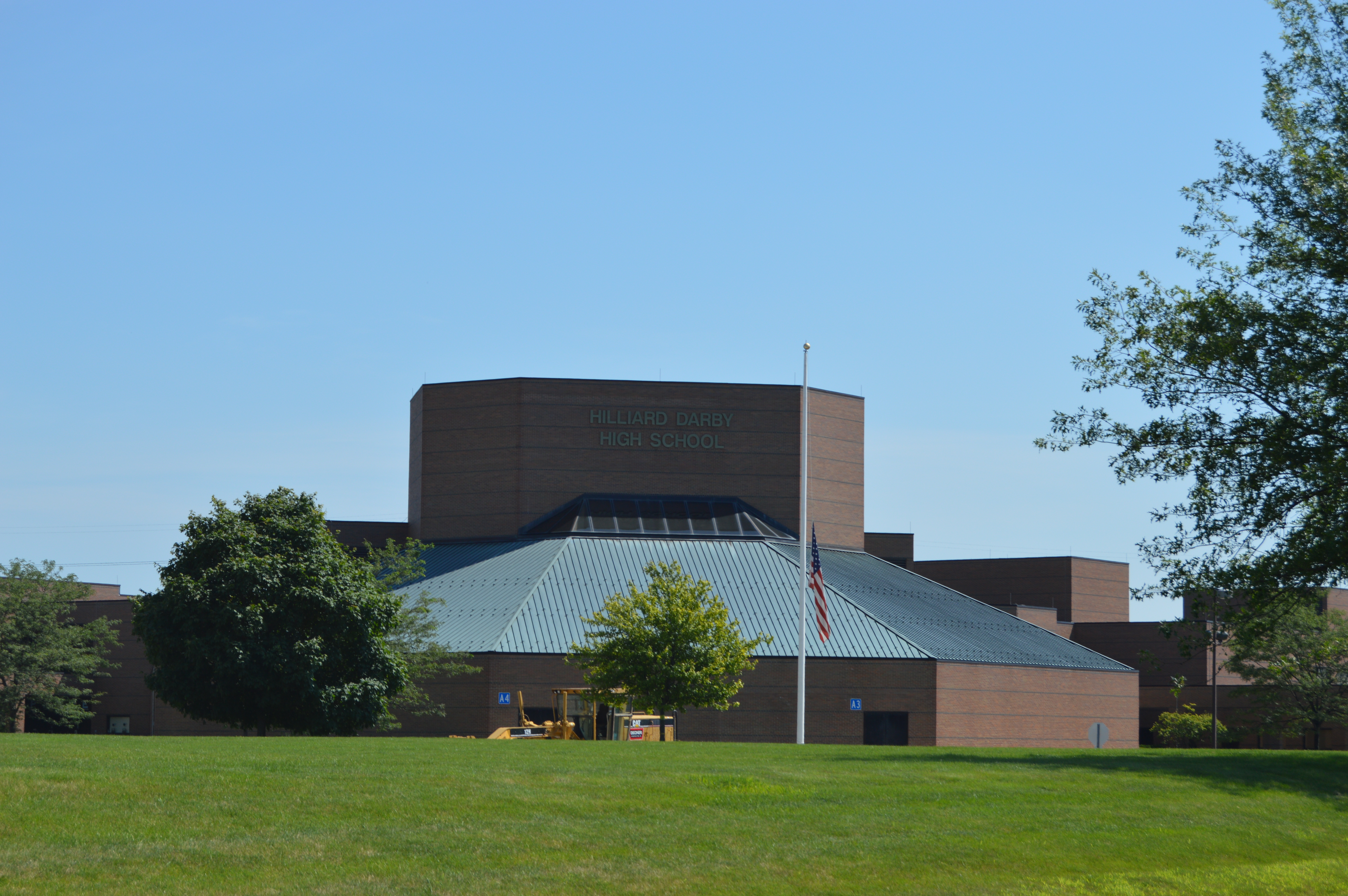 Brick high school building with a pyramid-shaped metal entrance roof and flagpole on a sunny day.
