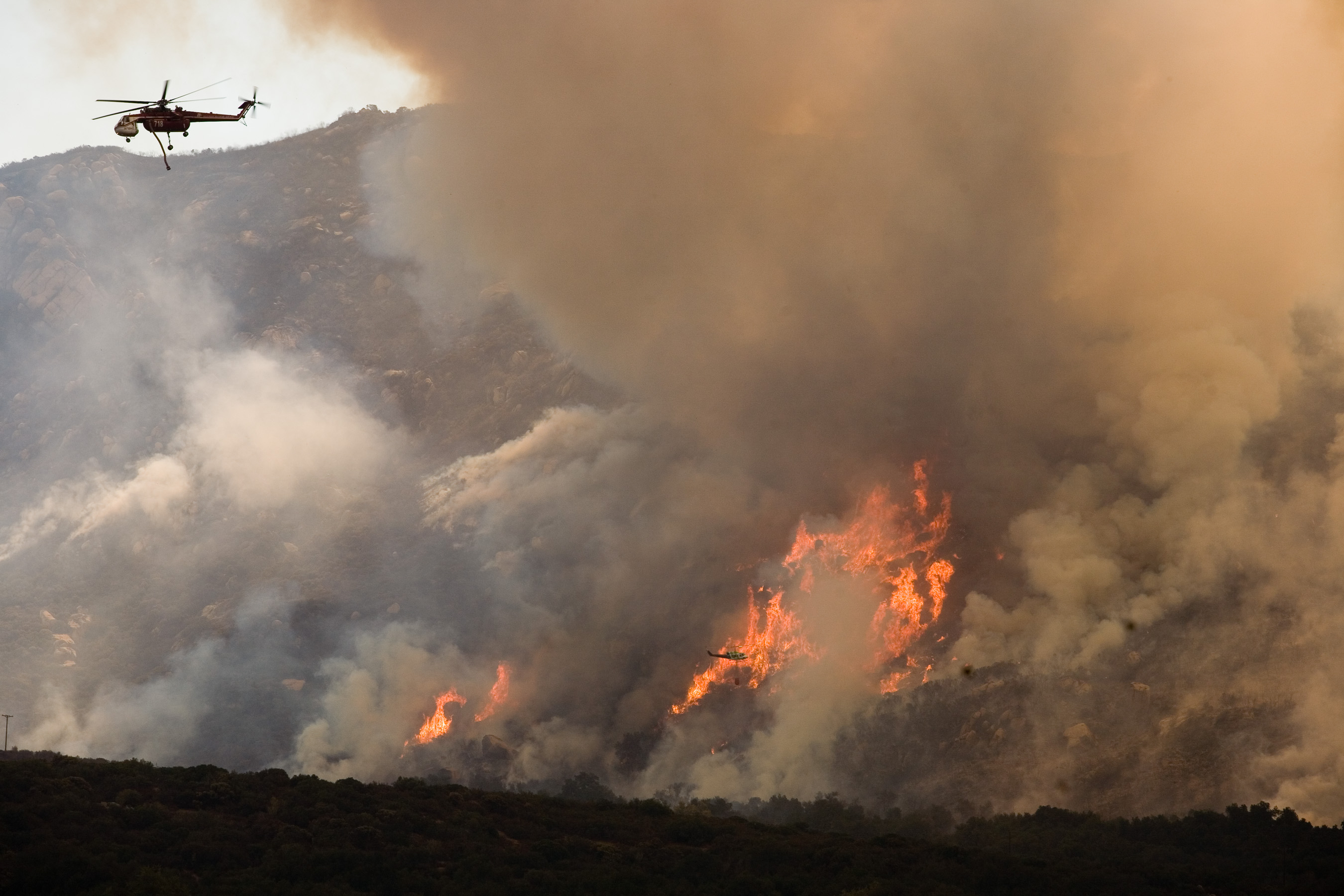 Helicopter drops retardant on a large wildfire burning on a brush-covered hillside.