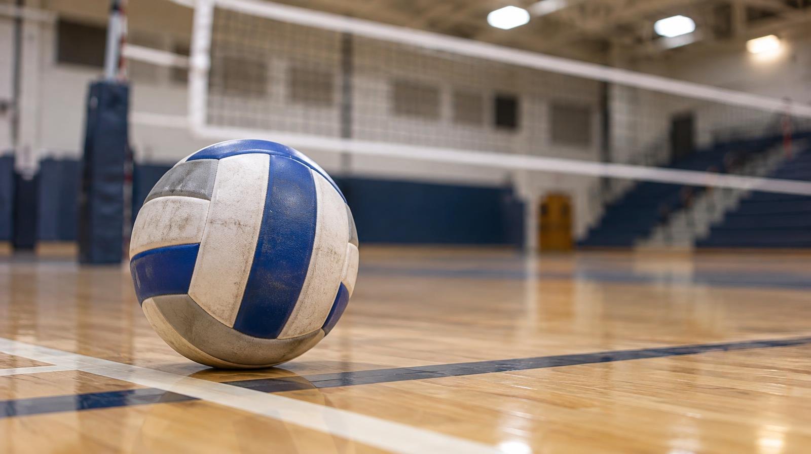 A volleyball sitting on a hardwood gym floor beside a net.