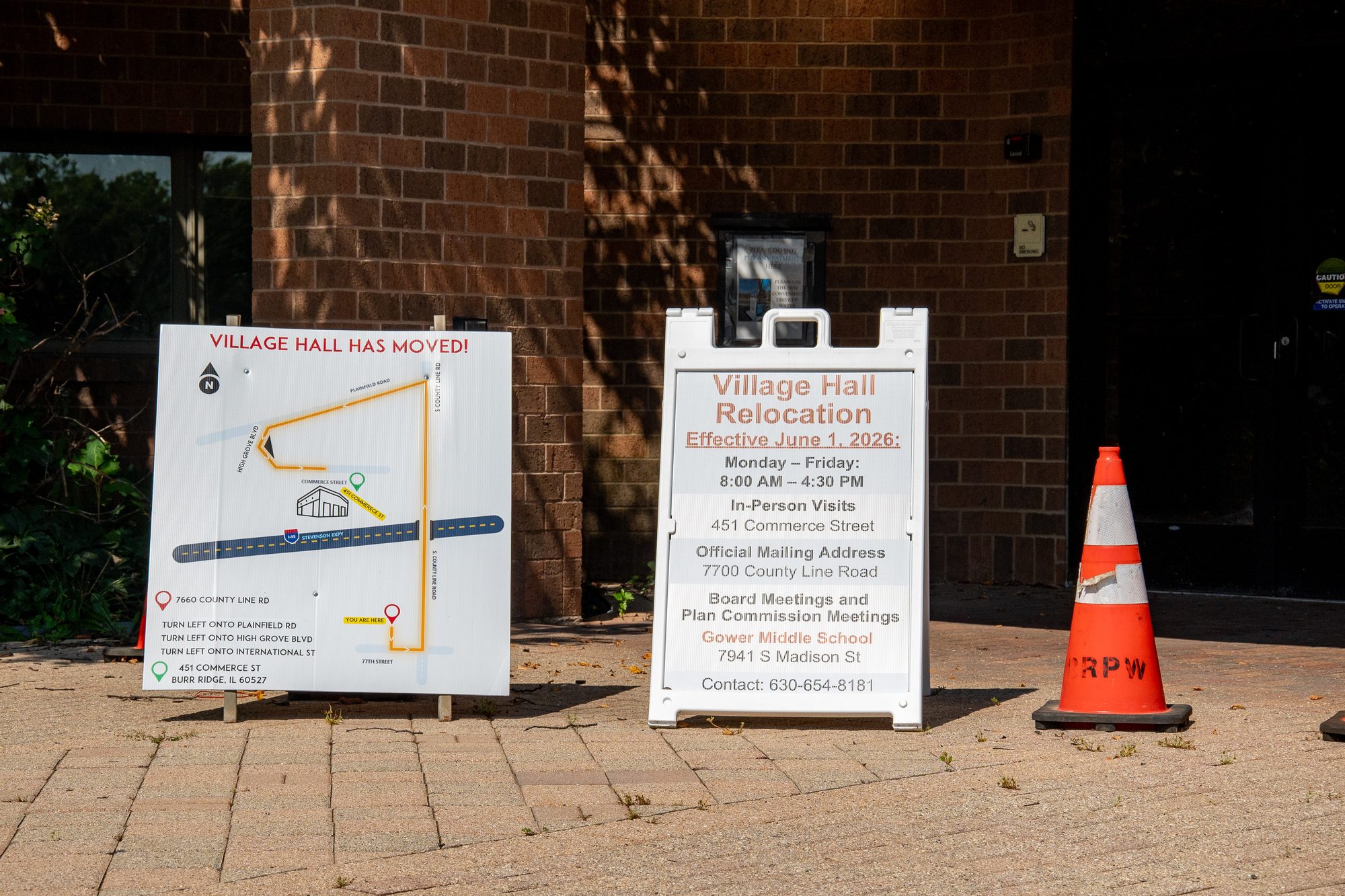 Two sandwich board signs outside a brick municipal building announcing a village hall relocation.