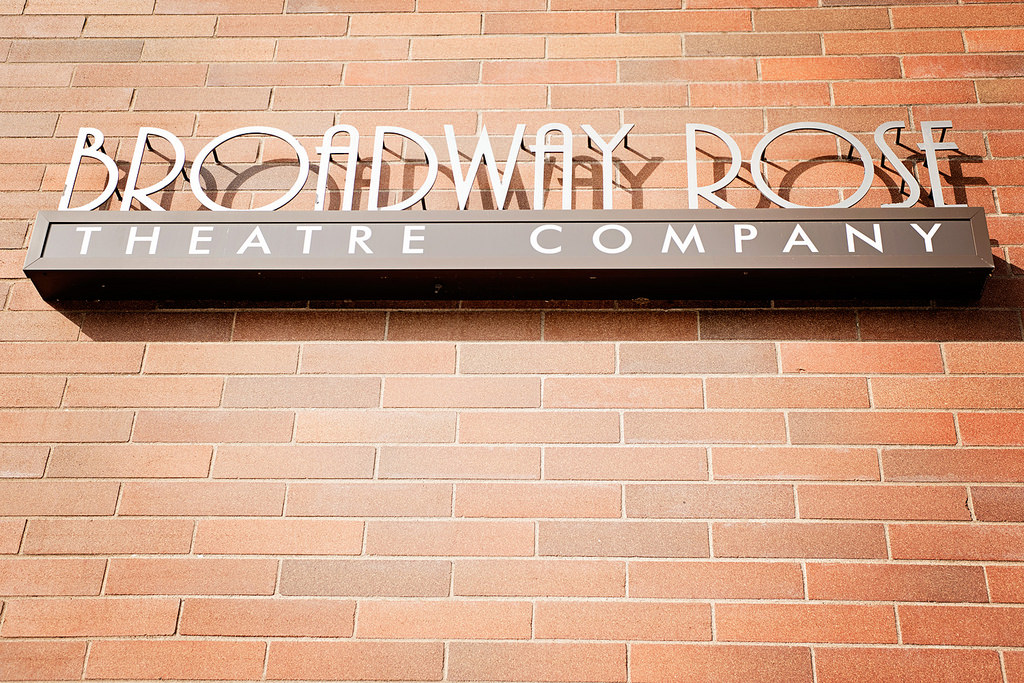 Sign reading Broadway Rose Theatre Company mounted on a red brick wall.