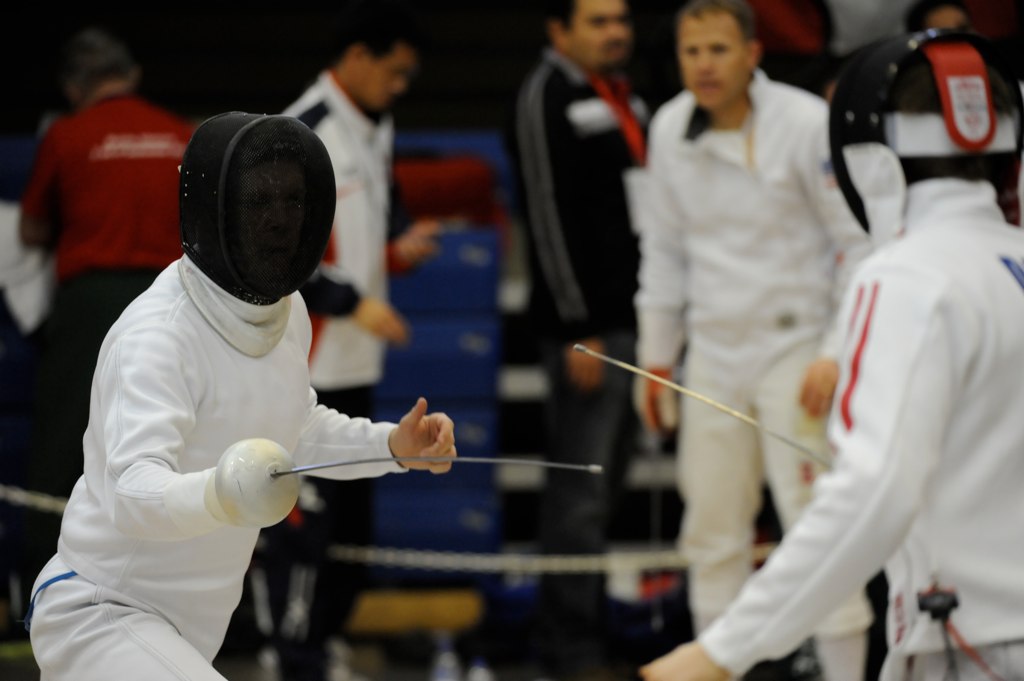 Two fencers in white jackets and protective masks square off indoors.