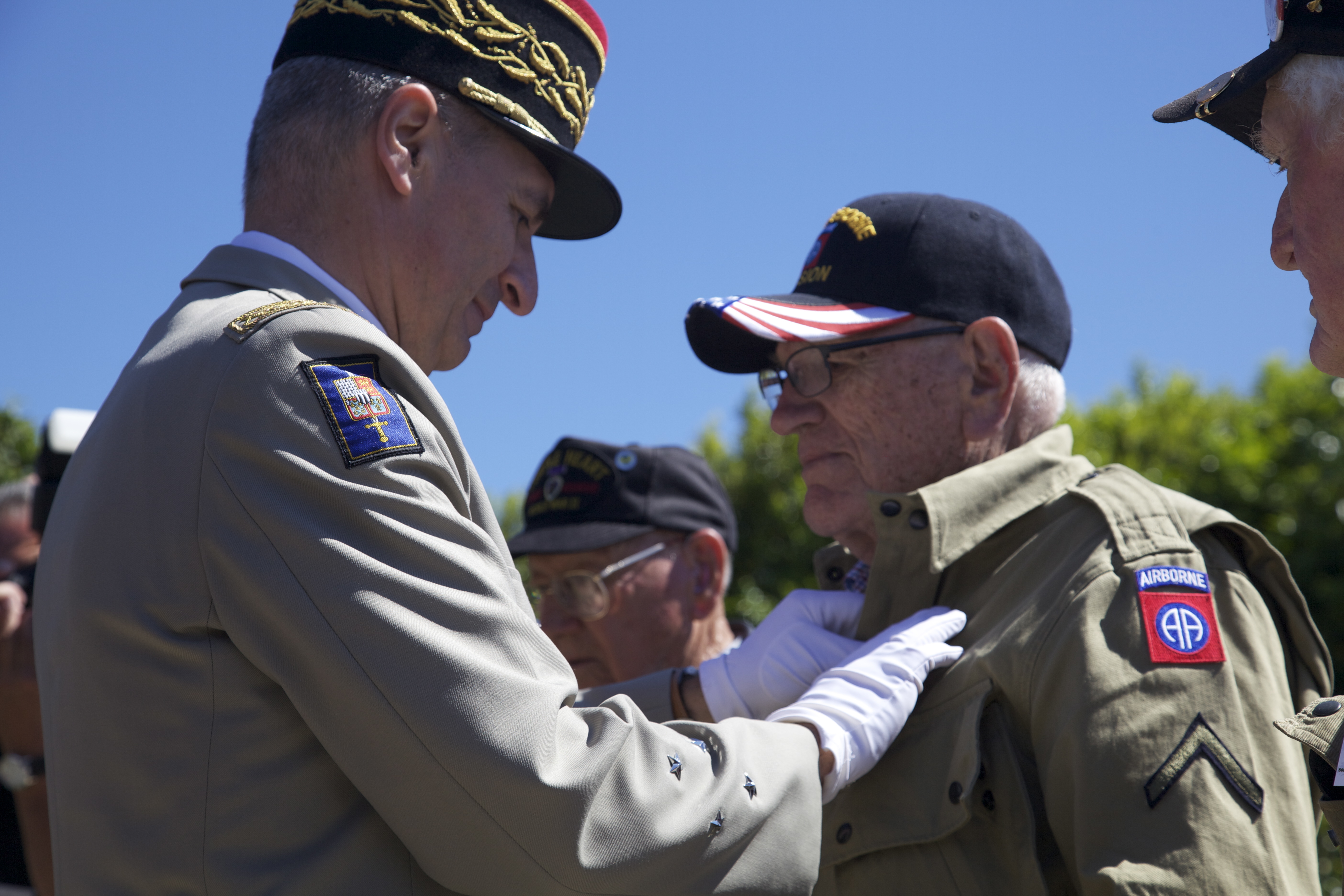 A military officer in white gloves pins a medal on an elderly veteran at an outdoor ceremony.