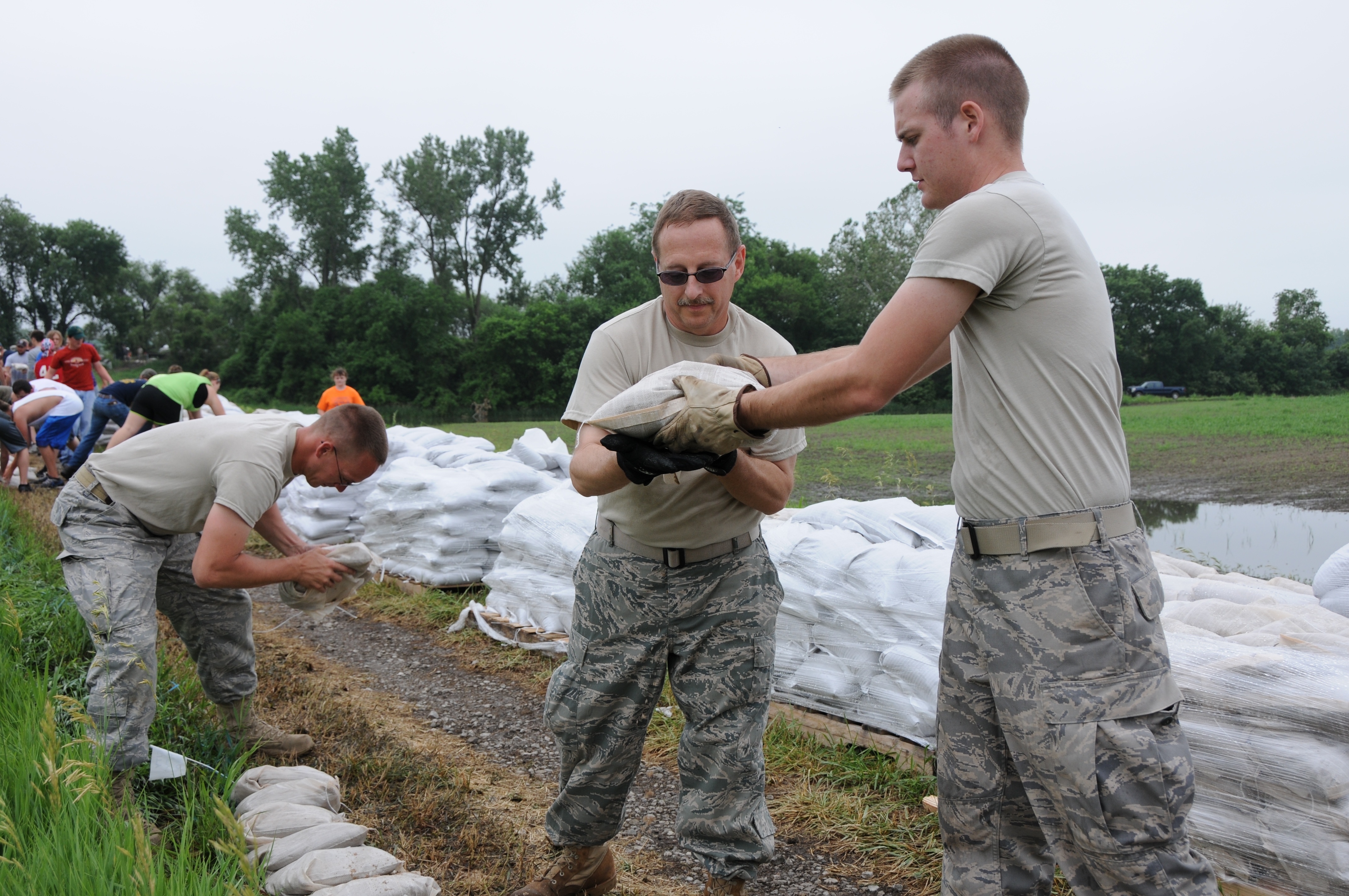 Three soldiers in camouflage uniforms stack white sandbags along a levee.