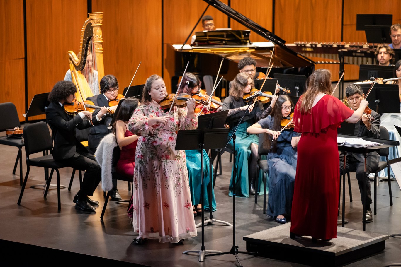 A violinist in a floral dress performs on stage while a conductor in a red dress leads an orchestra.