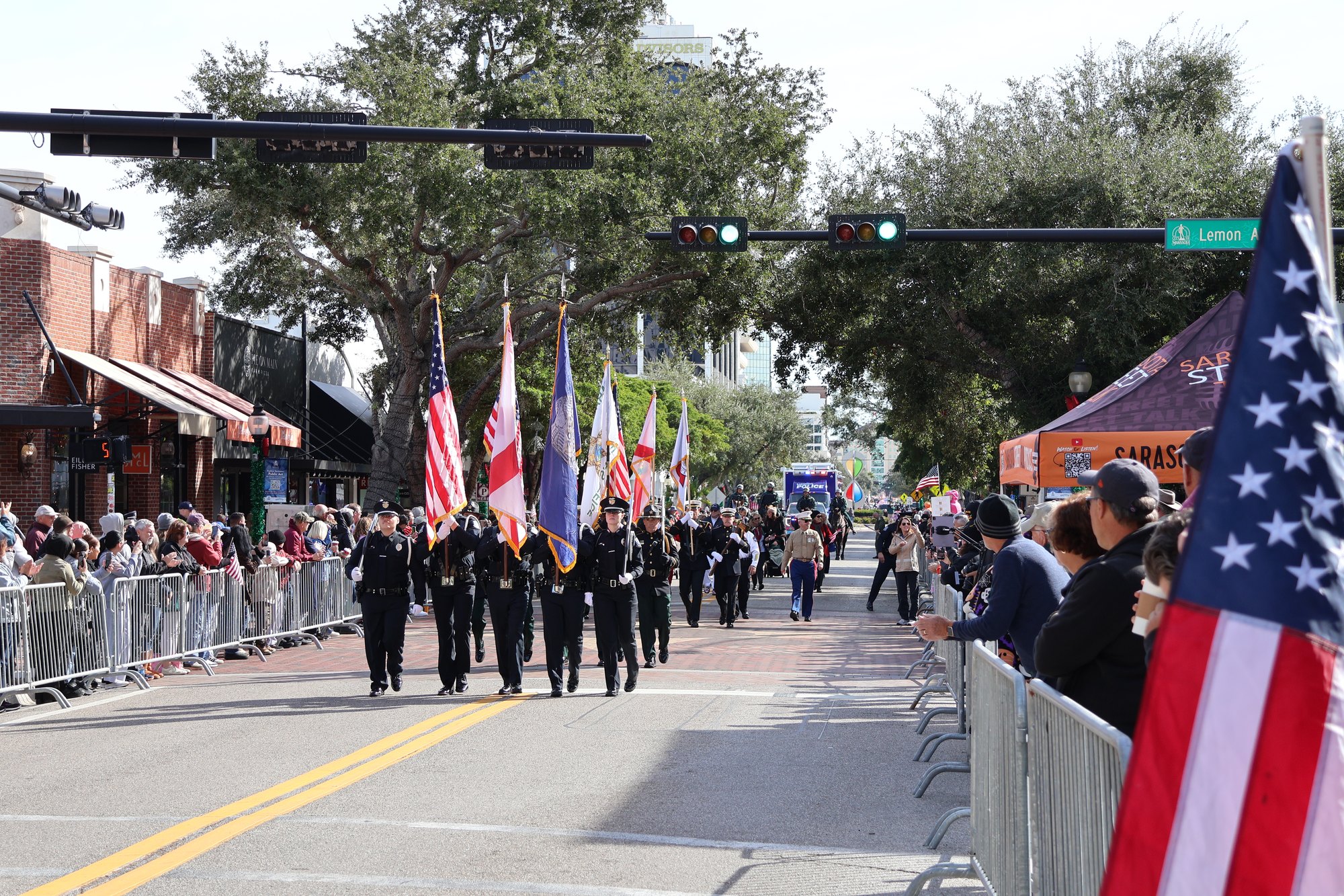 Uniformed color guard marches with American flags down a tree-lined city street.