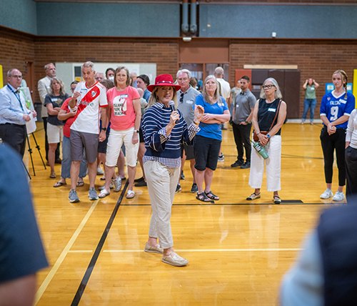 Crowd of adults gathered in a gymnasium around a woman in a red hat presenting at an easel.