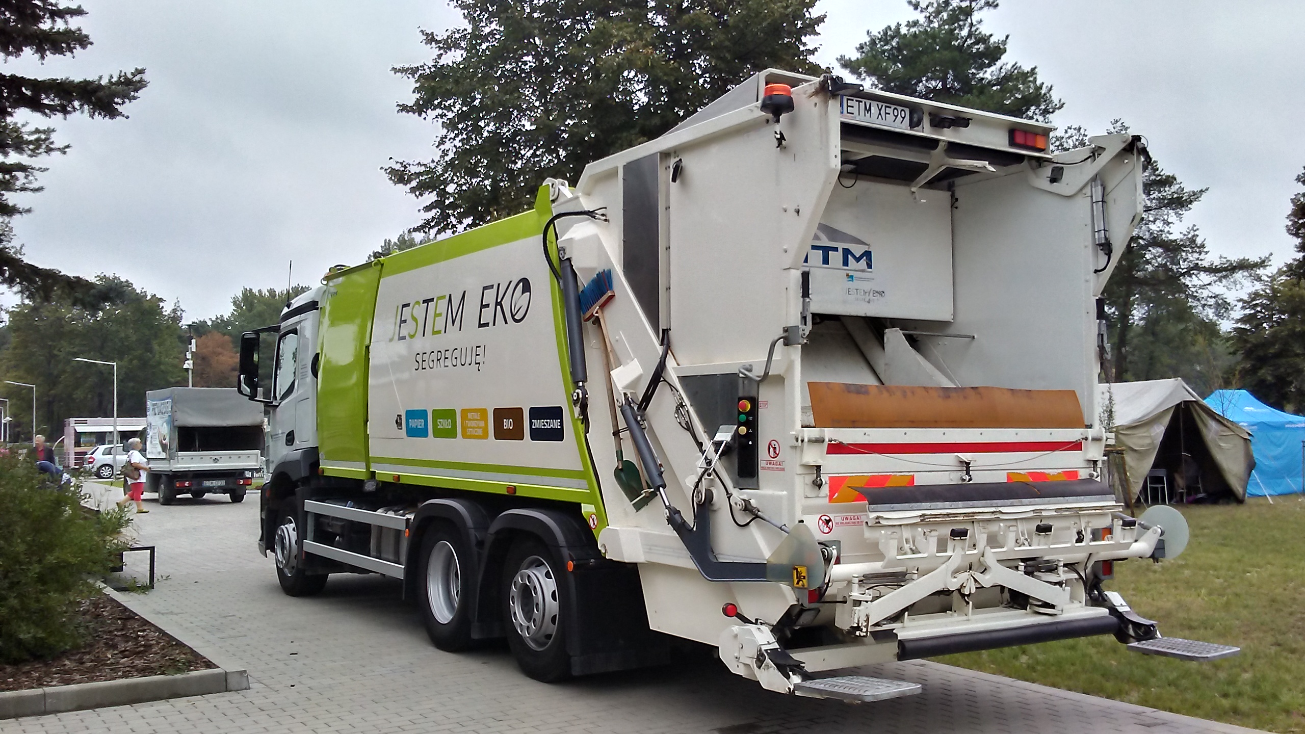 White and green rear-loading garbage truck parked on pavement near trees.