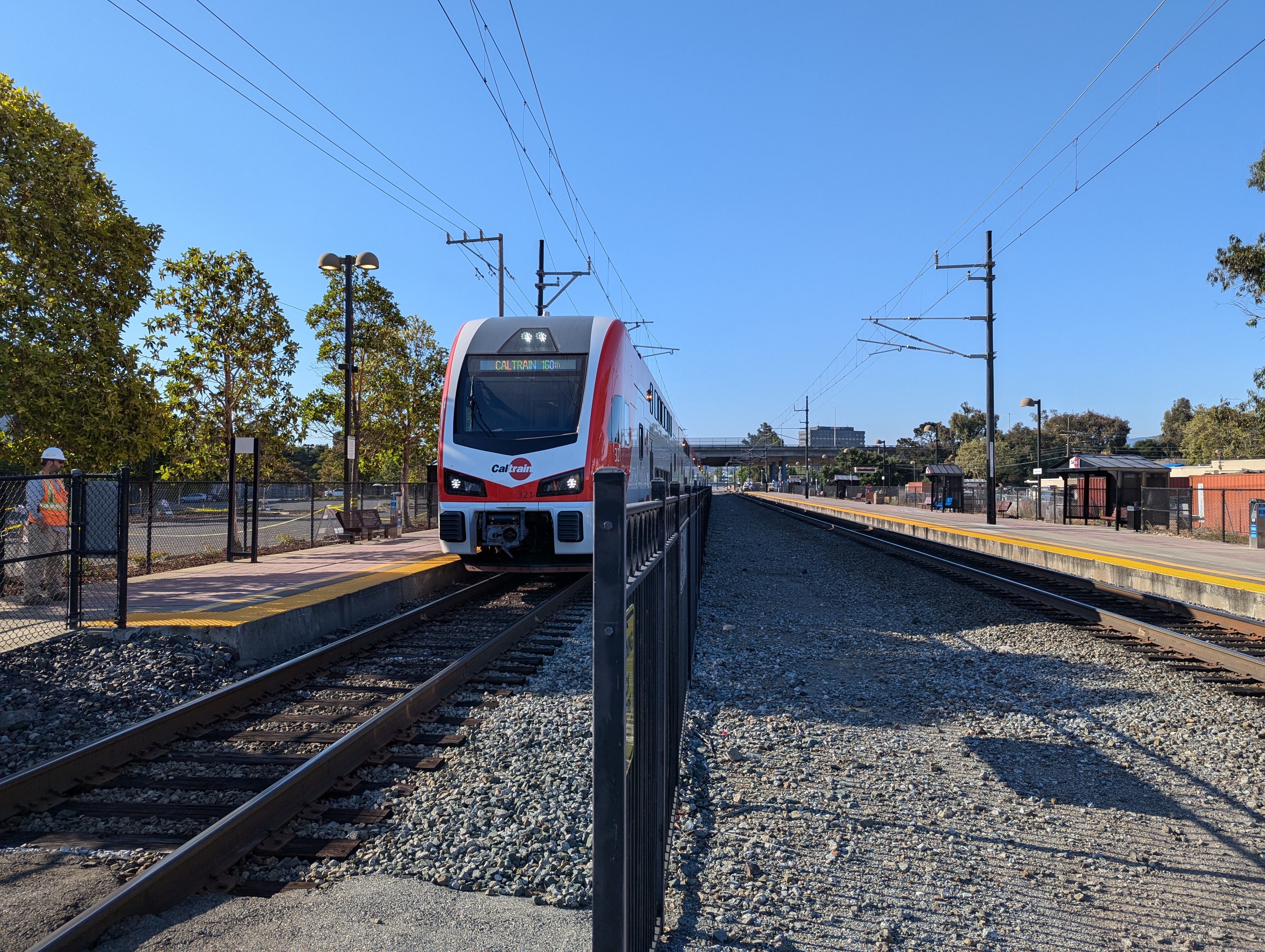 A Caltrain electric train approaching a station platform under clear blue skies with overhead wires.