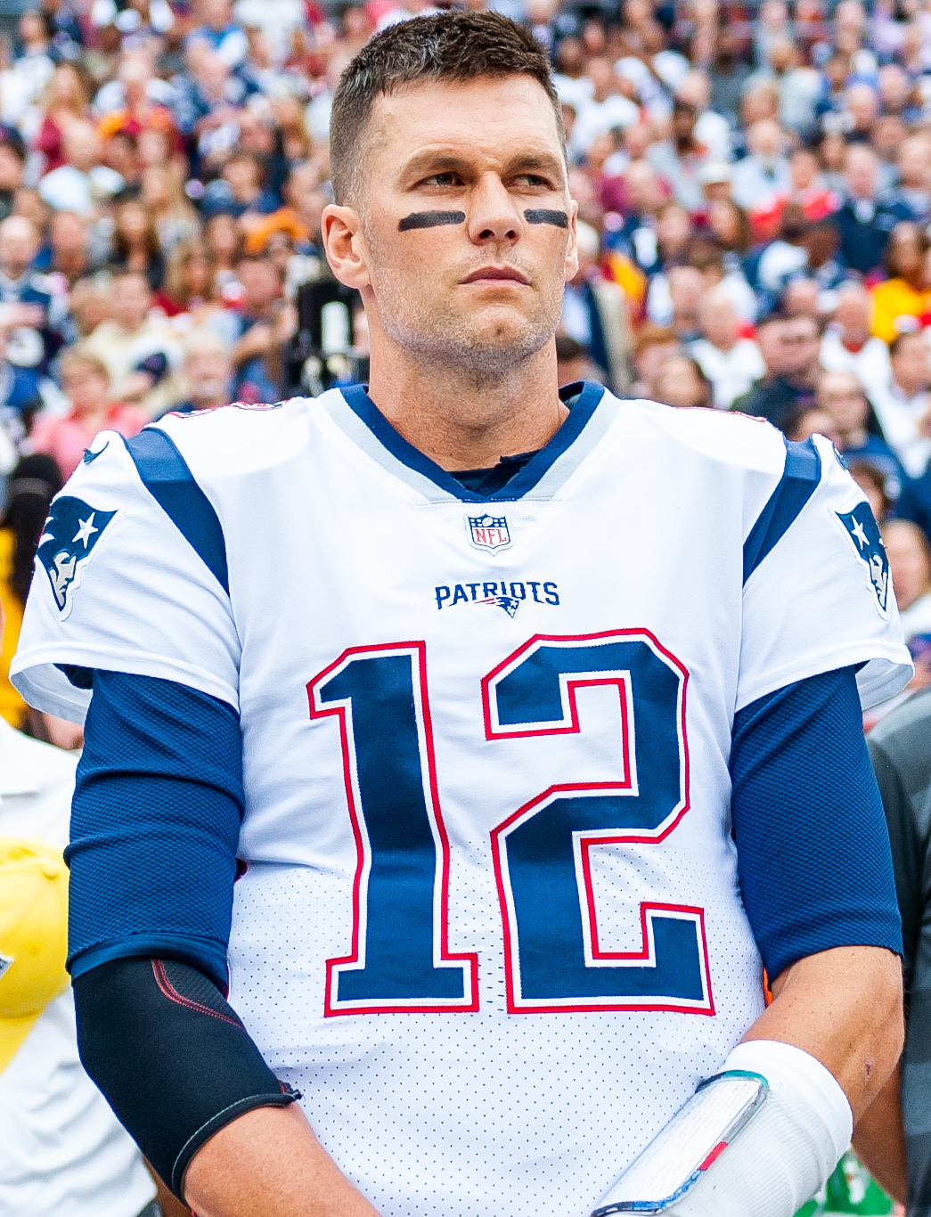 Tom Brady in a white New England Patriots jersey, number 12, standing on the sideline during a game.