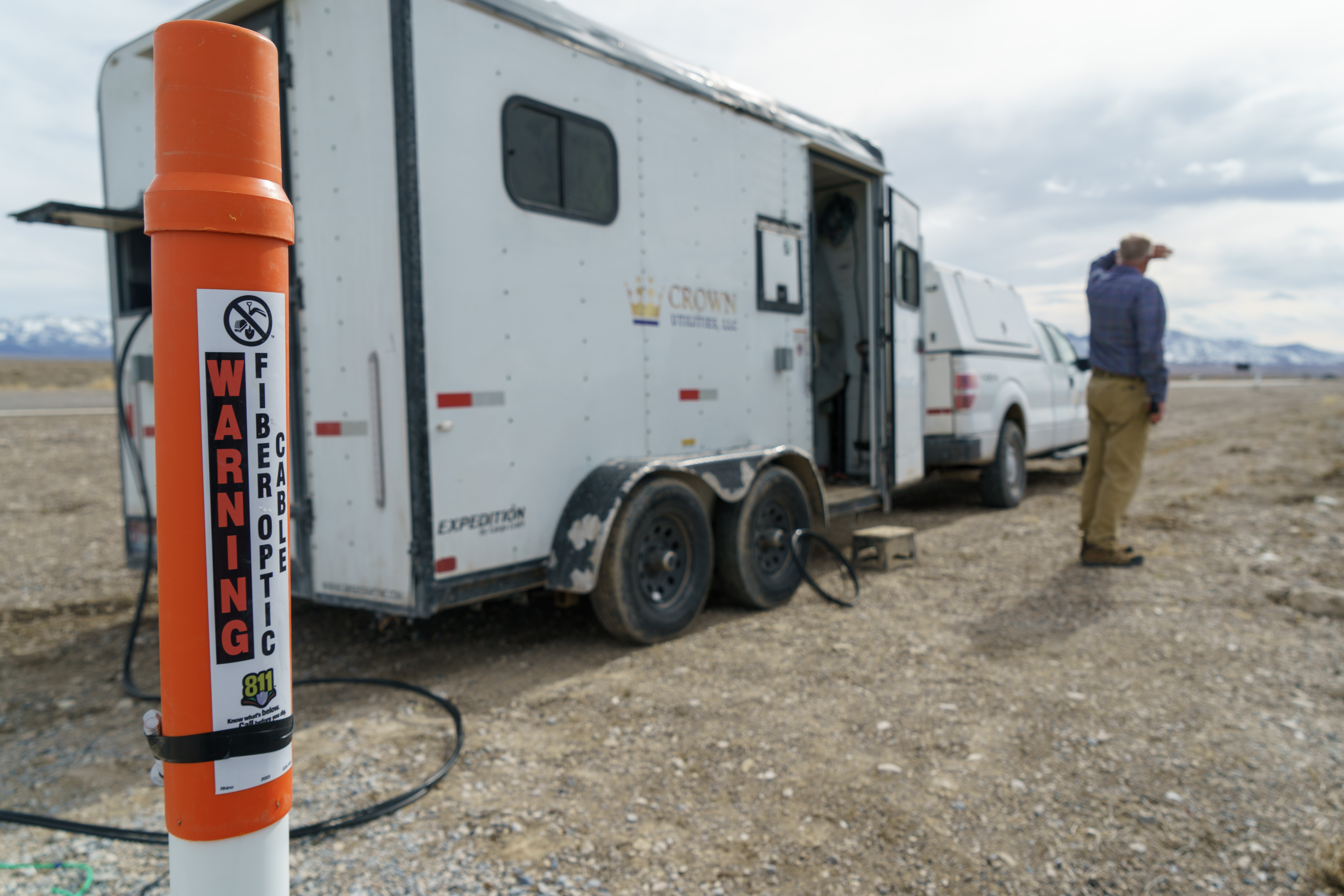 Orange fiber optic warning marker in foreground with utility trailer in a rural landscape.