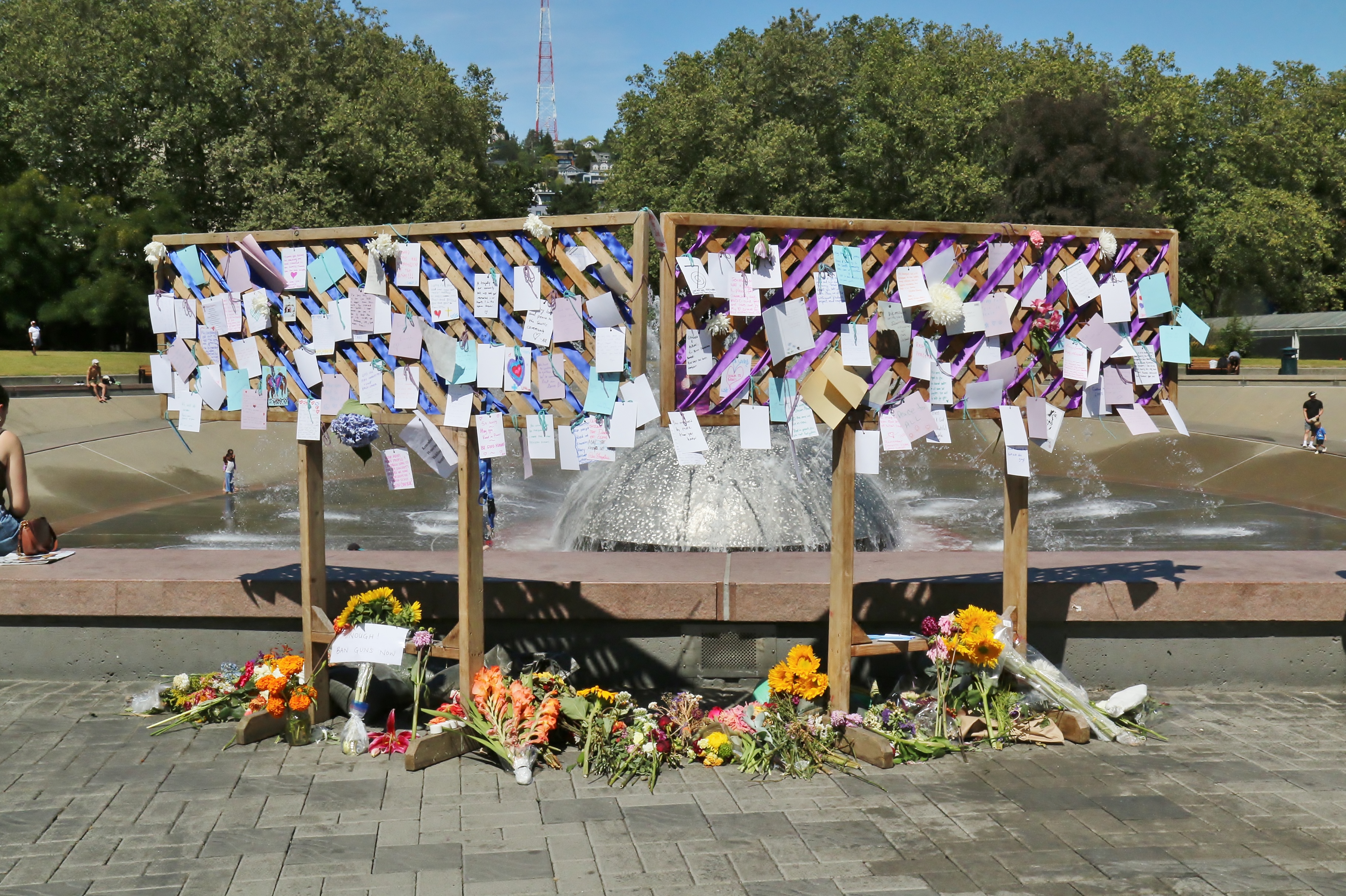 Wooden memorial panels covered in notes and purple ribbons stand before a fountain outdoors.
