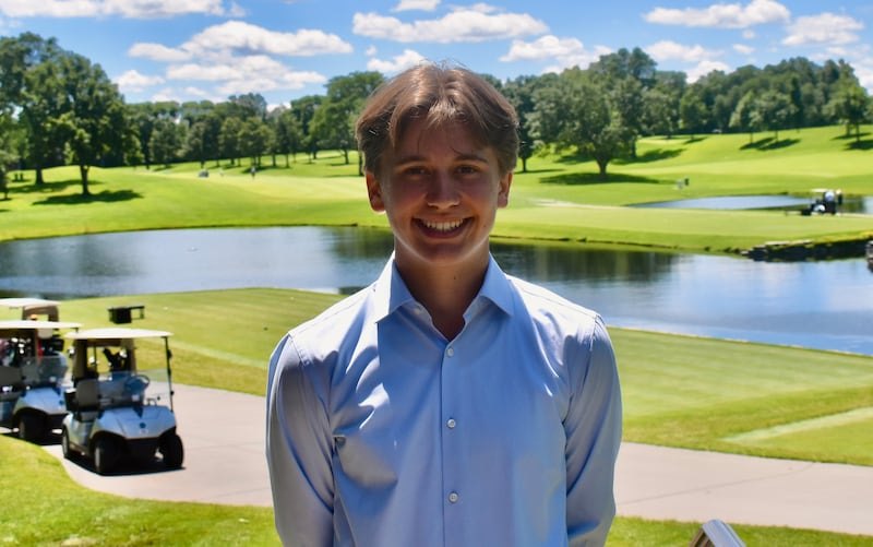 Young man in a light blue shirt smiling on a patio overlooking a golf course with a pond and golf carts.