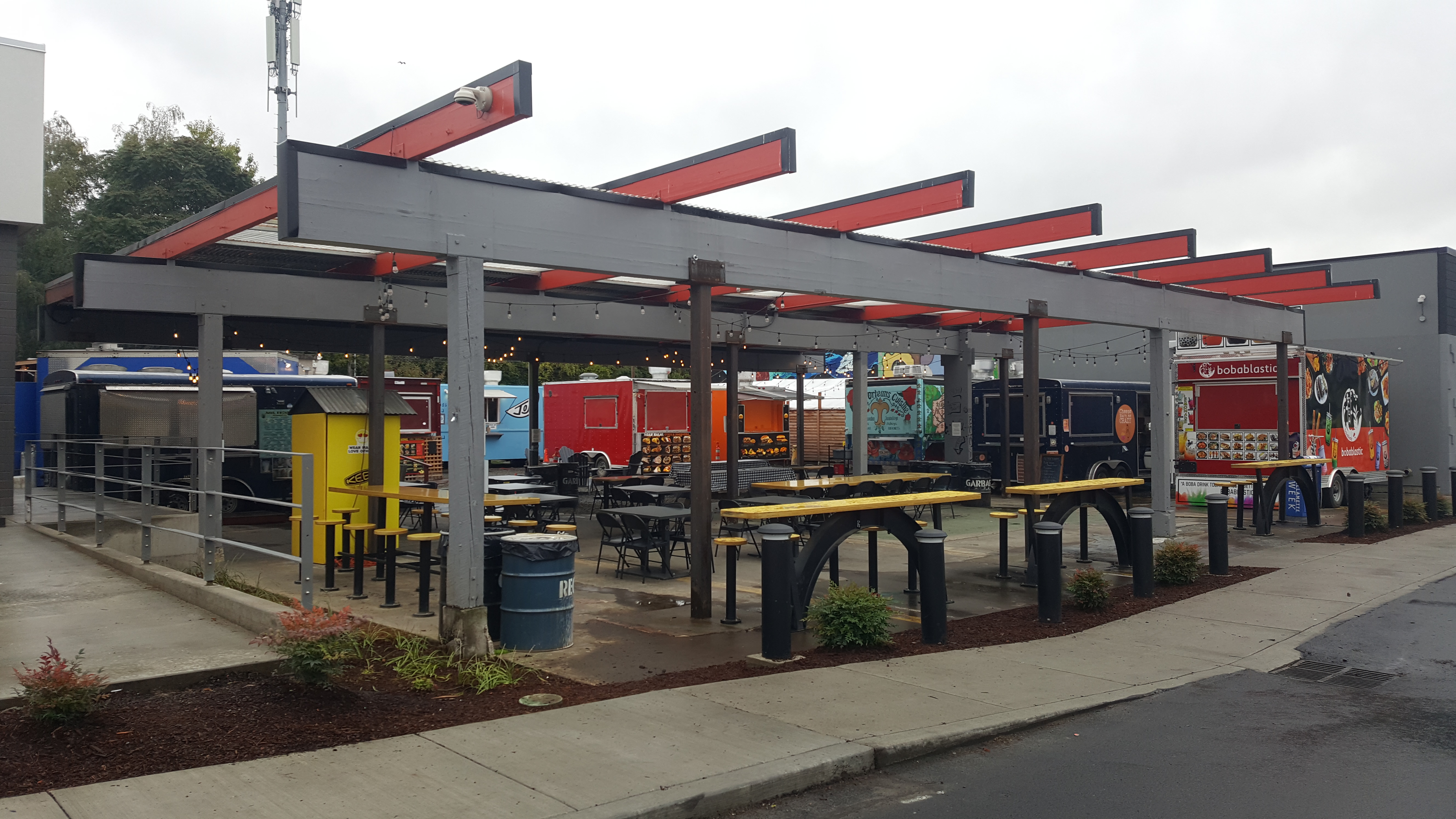 Covered outdoor food cart pod with metal pergola, picnic tables, and food trailers.