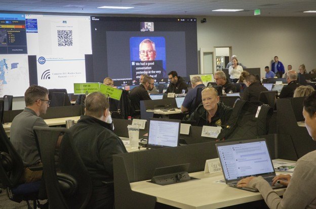 People seated at laptops in a large room with display screens at the front.
