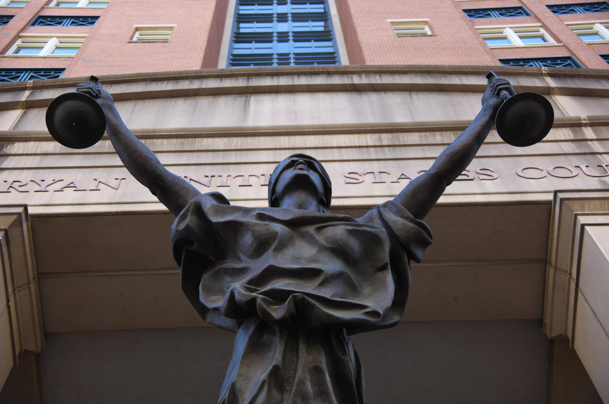 Bronze Blind Justice statue with outstretched scales photographed from below against a courthouse facade.