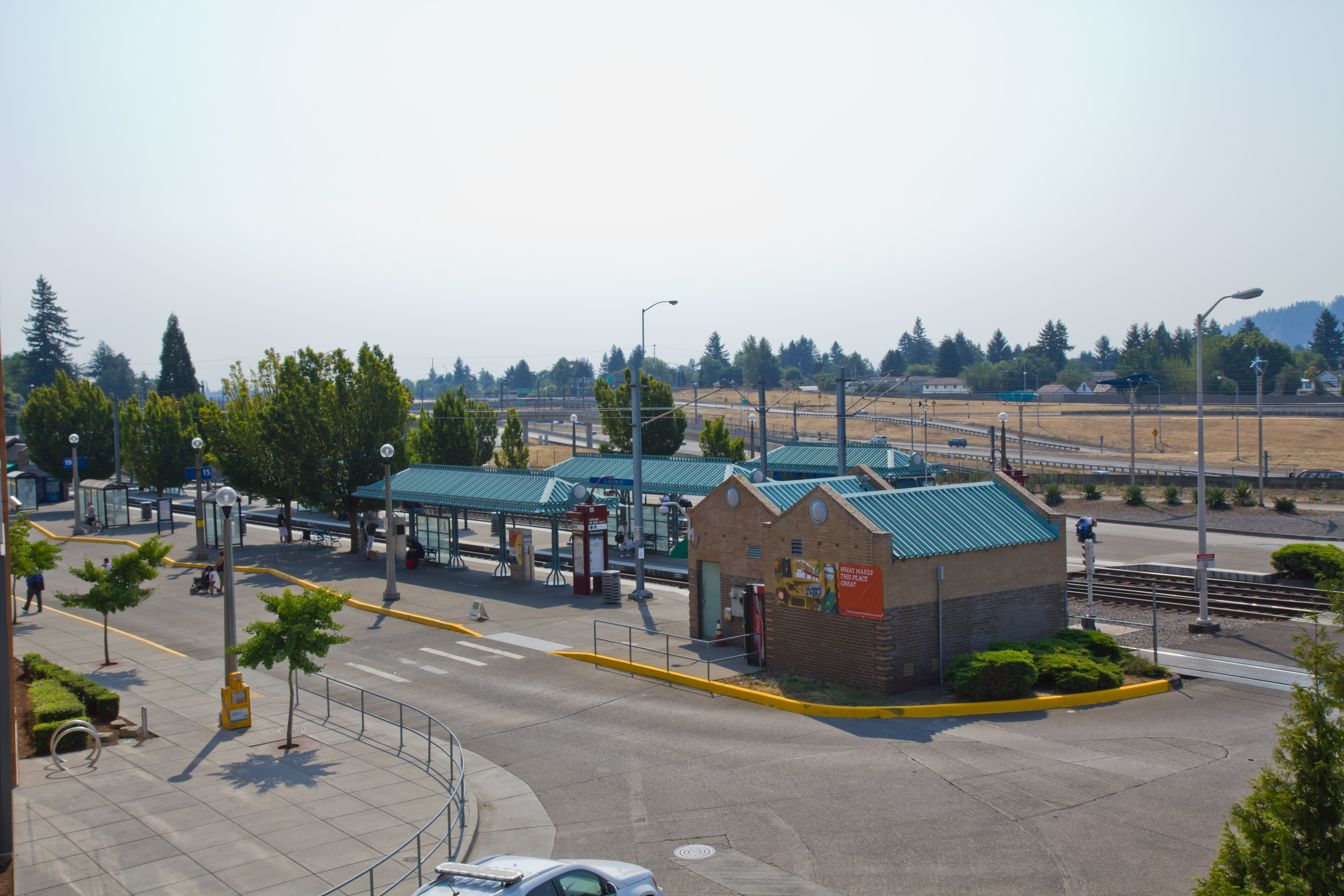 Elevated view of a transit center with covered bus shelters, a brick building, and light rail tracks.