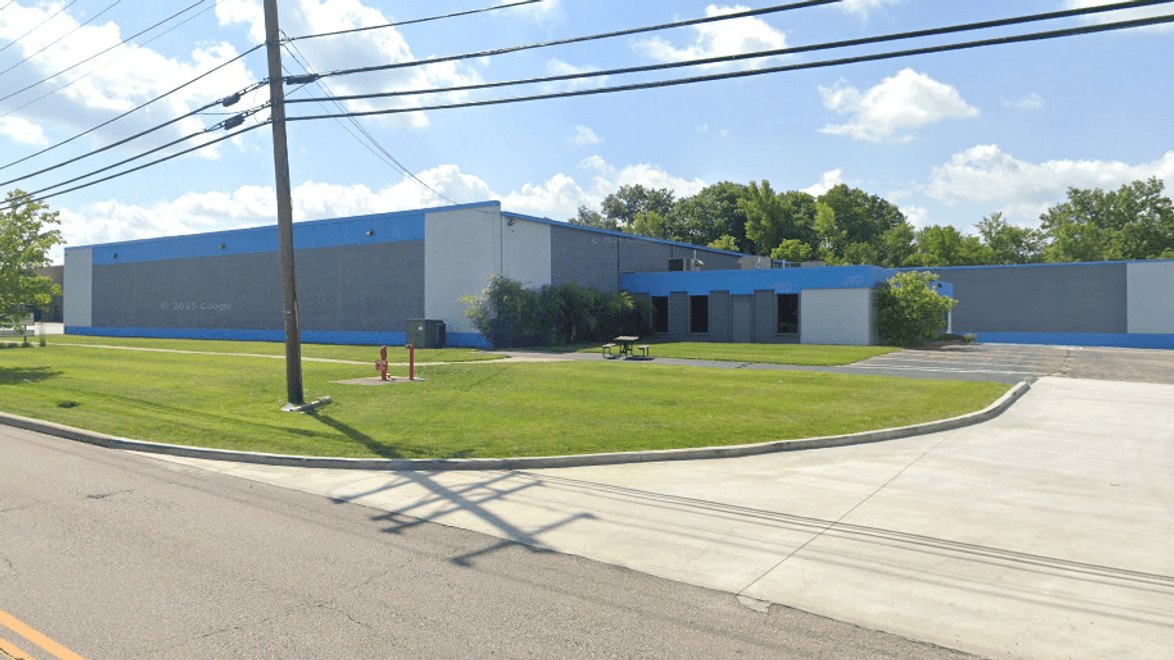 Large gray warehouse building with blue trim band, surrounded by green lawn and utility poles.