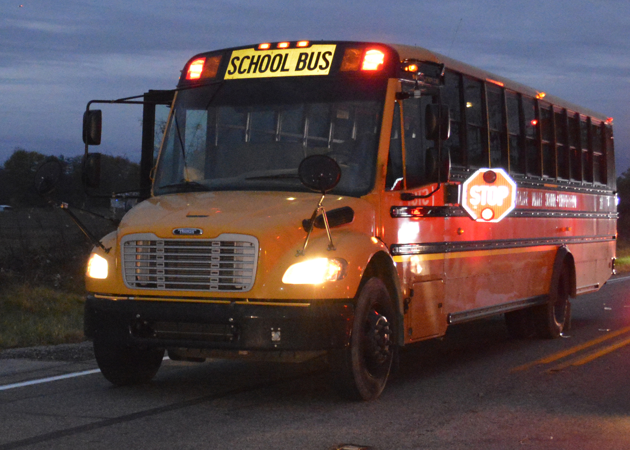 Yellow school bus on a road at dusk with red warning lights and stop arm extended.
