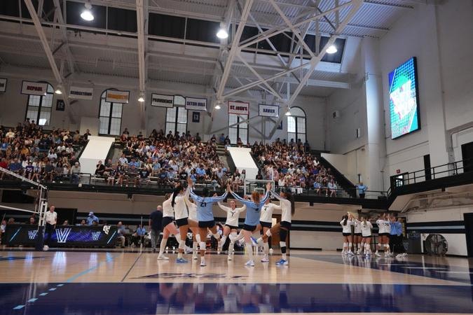 Women's volleyball team in light blue uniforms huddles on an indoor court.