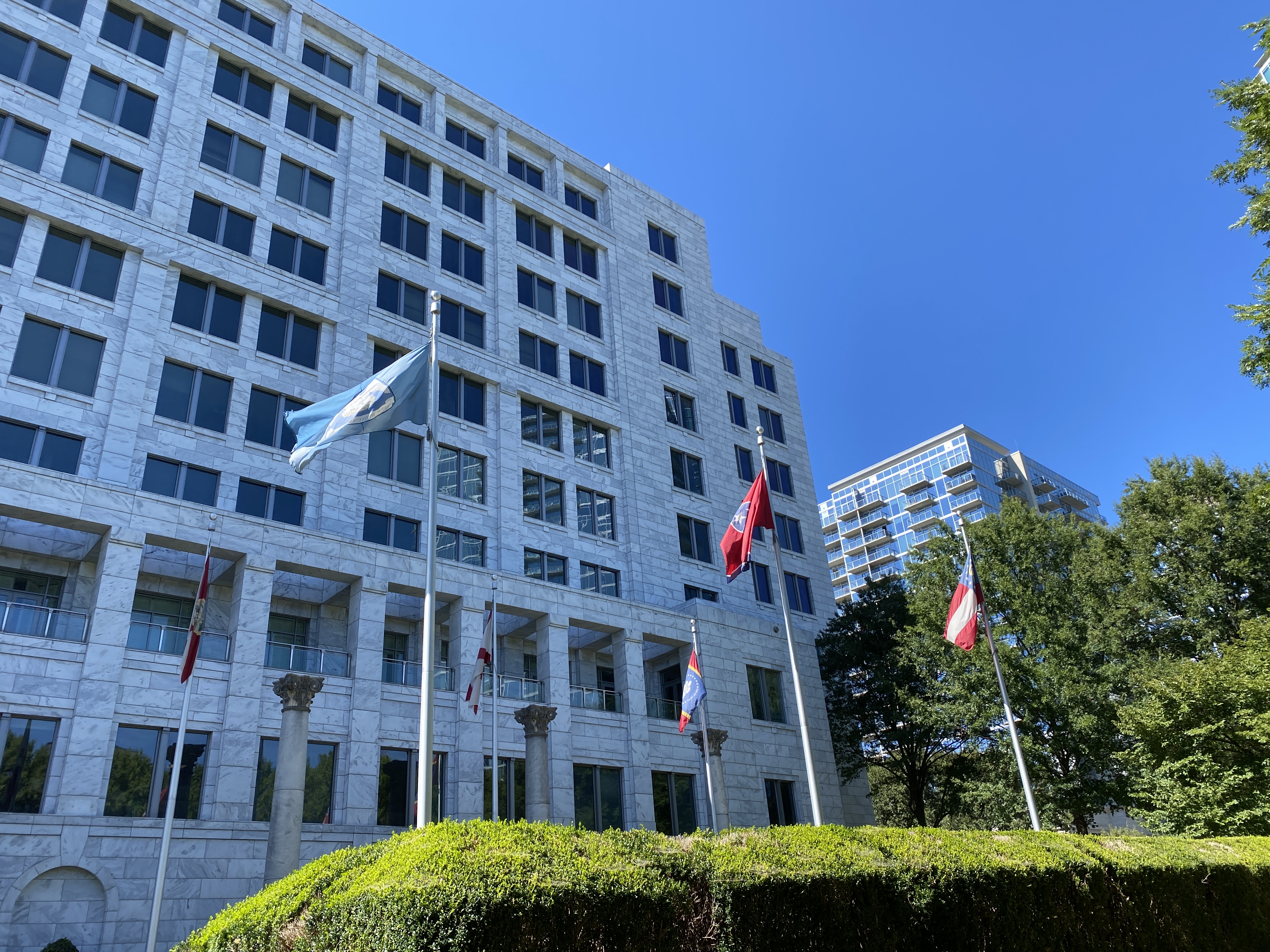 Light stone Federal Reserve Bank building with multiple flags and hedges under a clear blue sky.
