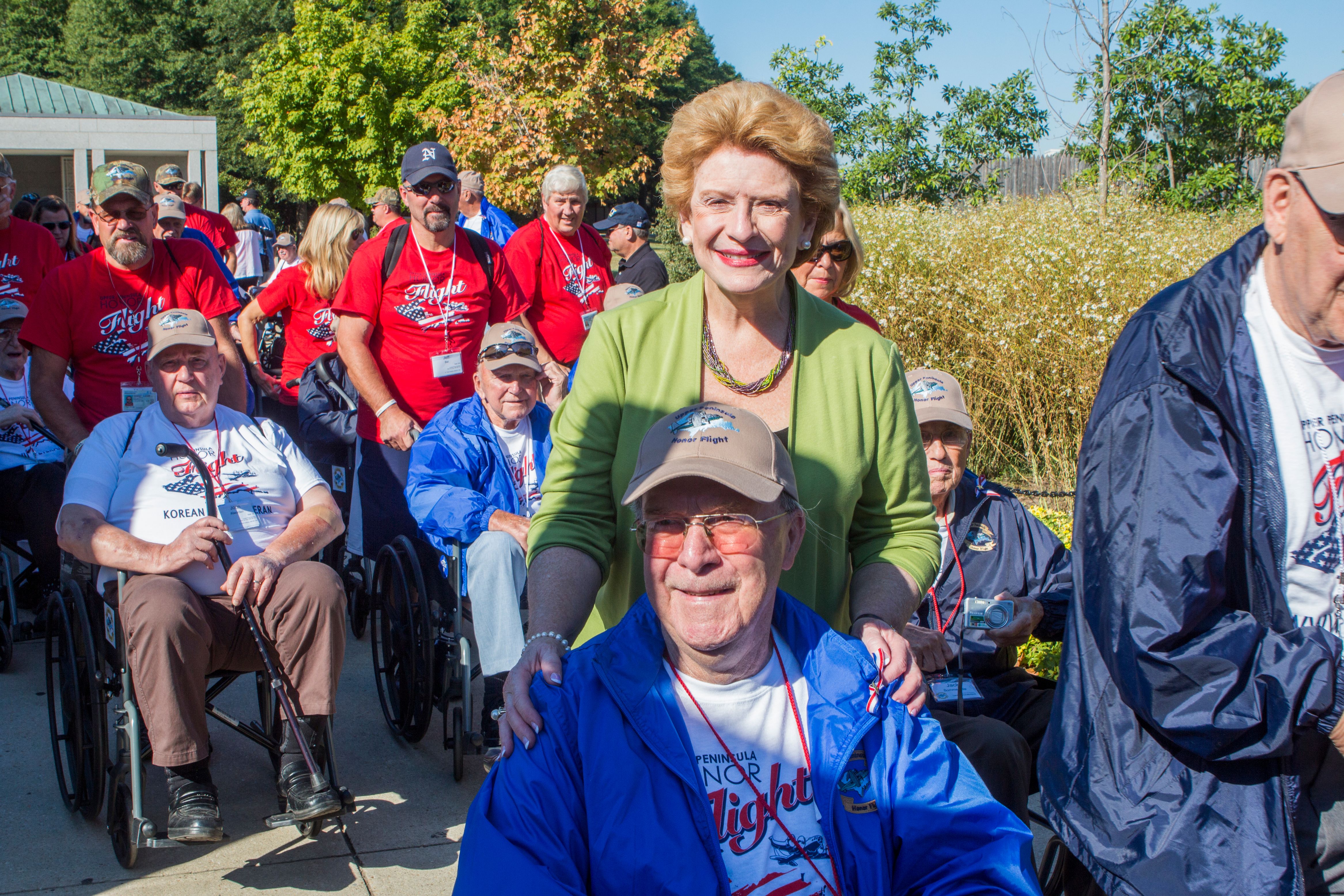 Elderly veterans in wheelchairs wearing red Honor Flight shirts at an outdoor memorial.