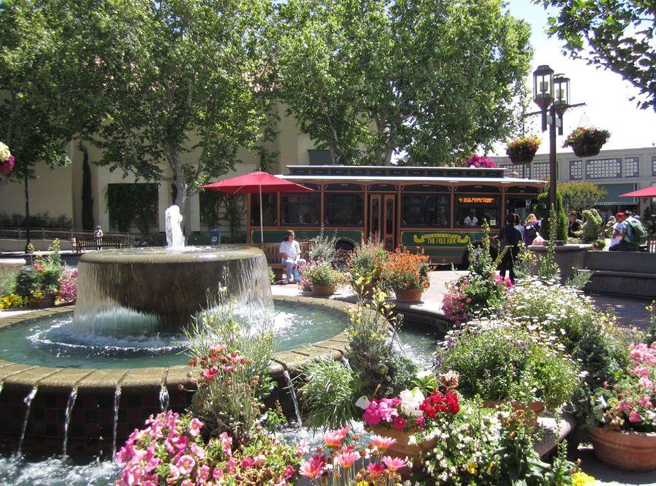Outdoor shopping plaza courtyard with a tiered stone fountain, flower planters, and shoppers walking past.