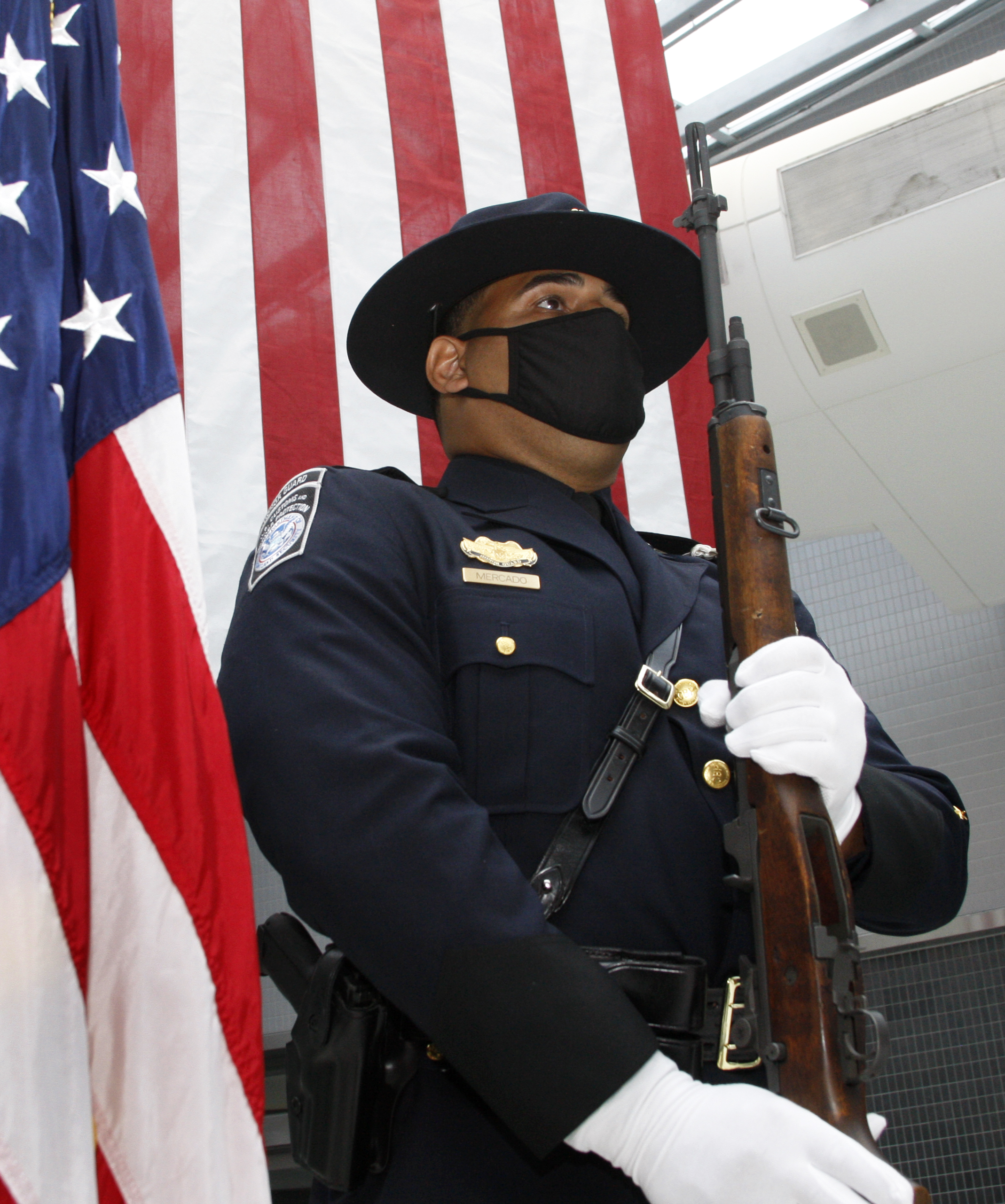 Uniformed officer in campaign hat stands at attention holding a rifle before an American flag.