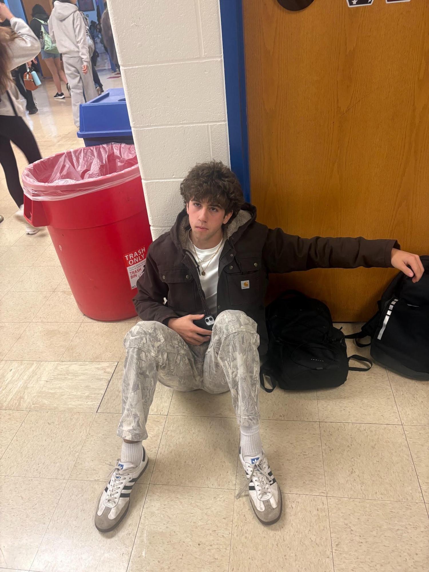 A student sitting on a school hallway floor next to a backpack and a red trash can.
