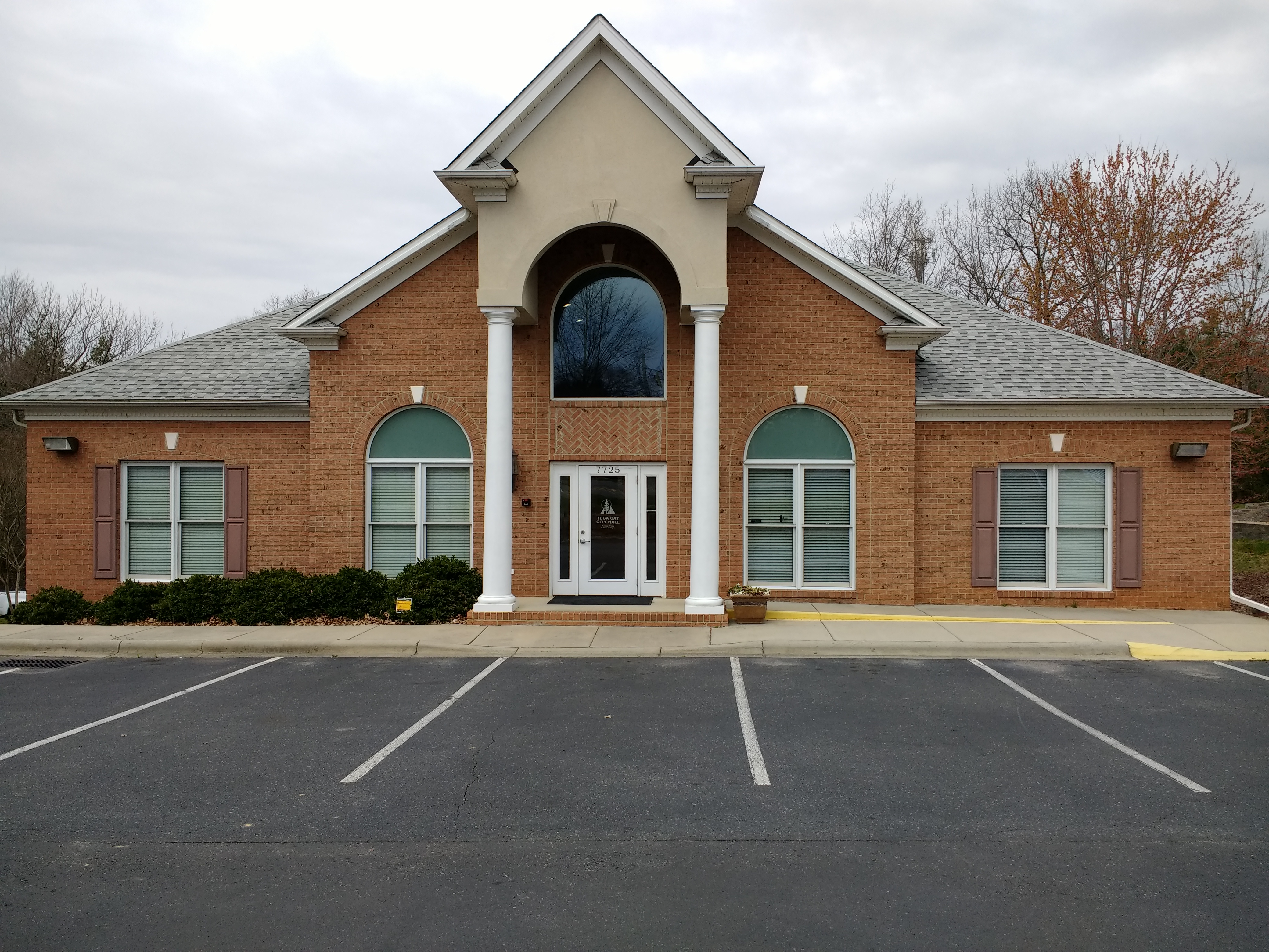 Brick civic building with white columns and arched windows, empty parking lot in front.
