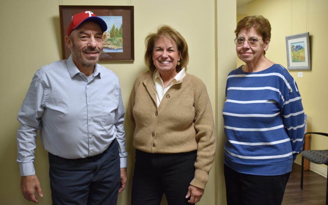 Two women and a man wearing a red Phillies cap smile together indoors at a community center.