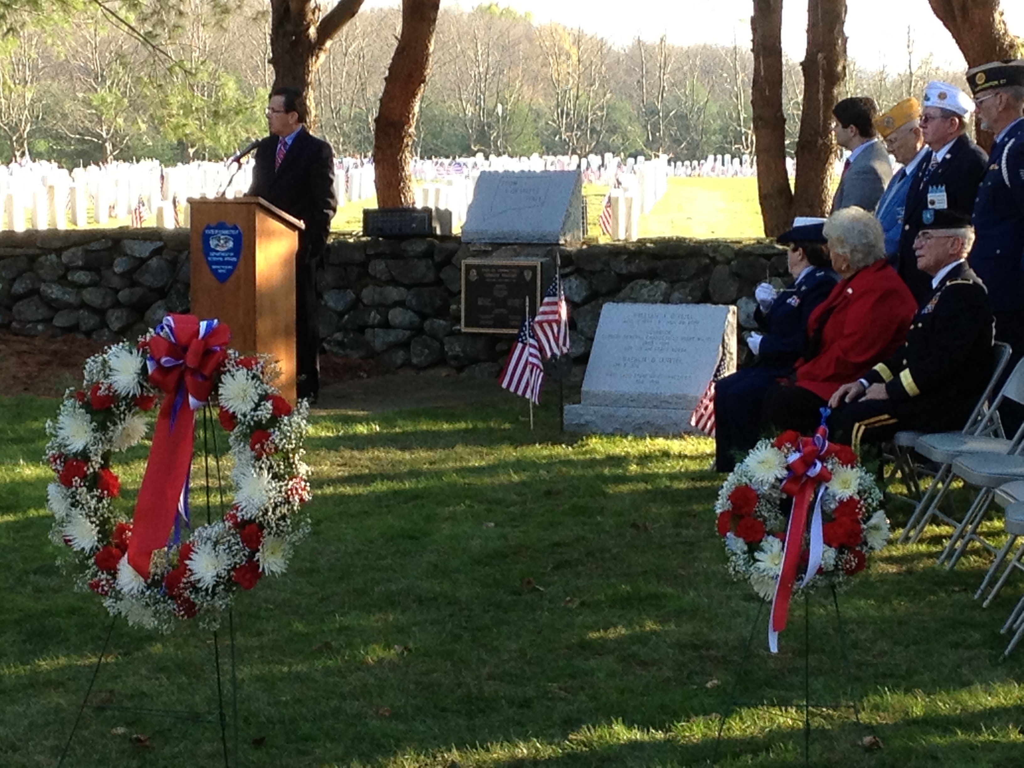 Speaker at a podium flanked by two red, white, and blue wreaths at an outdoor Veterans Day ceremony.