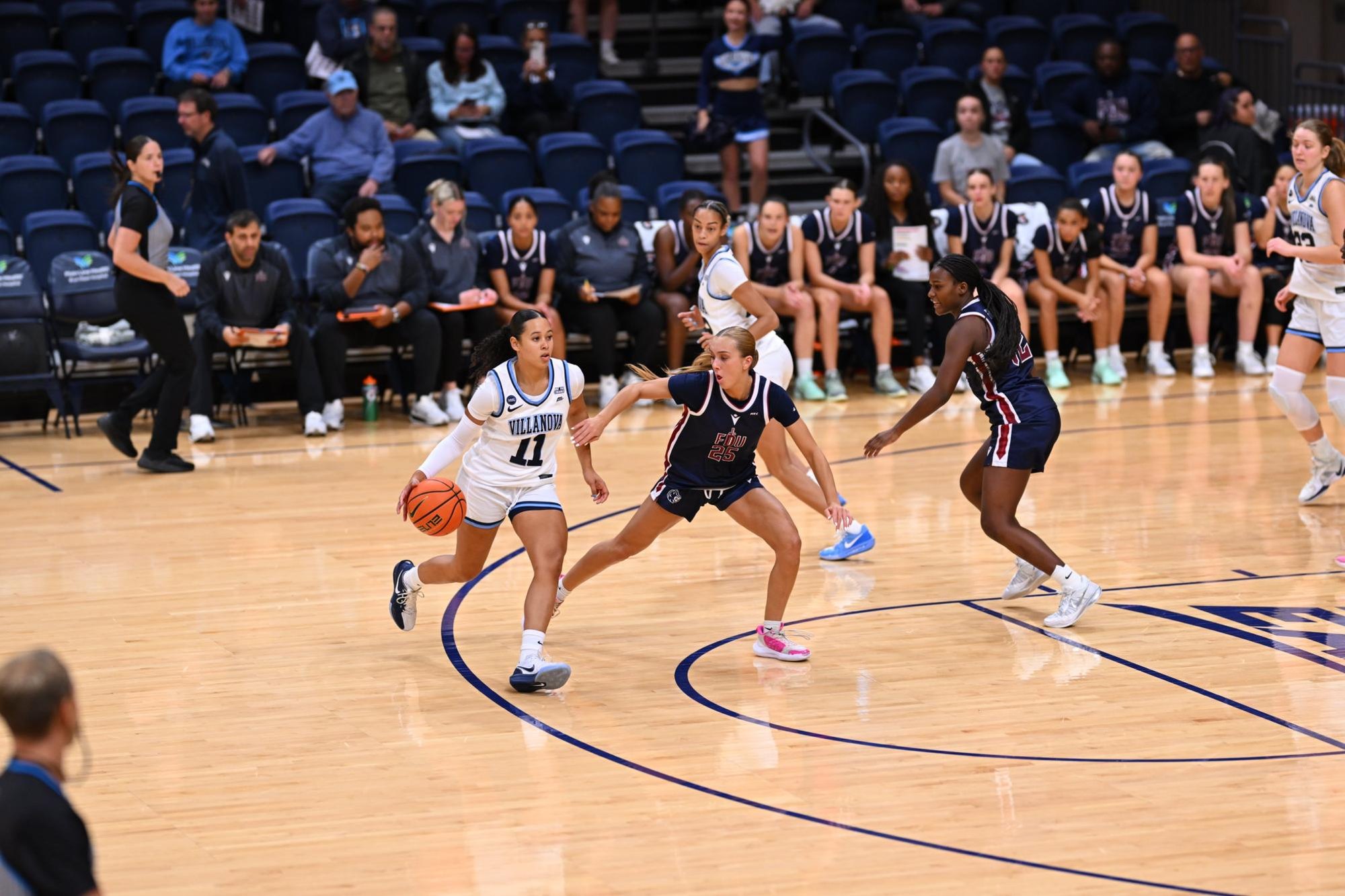 A Villanova women's basketball player in white dribbles past two defenders in dark uniforms.